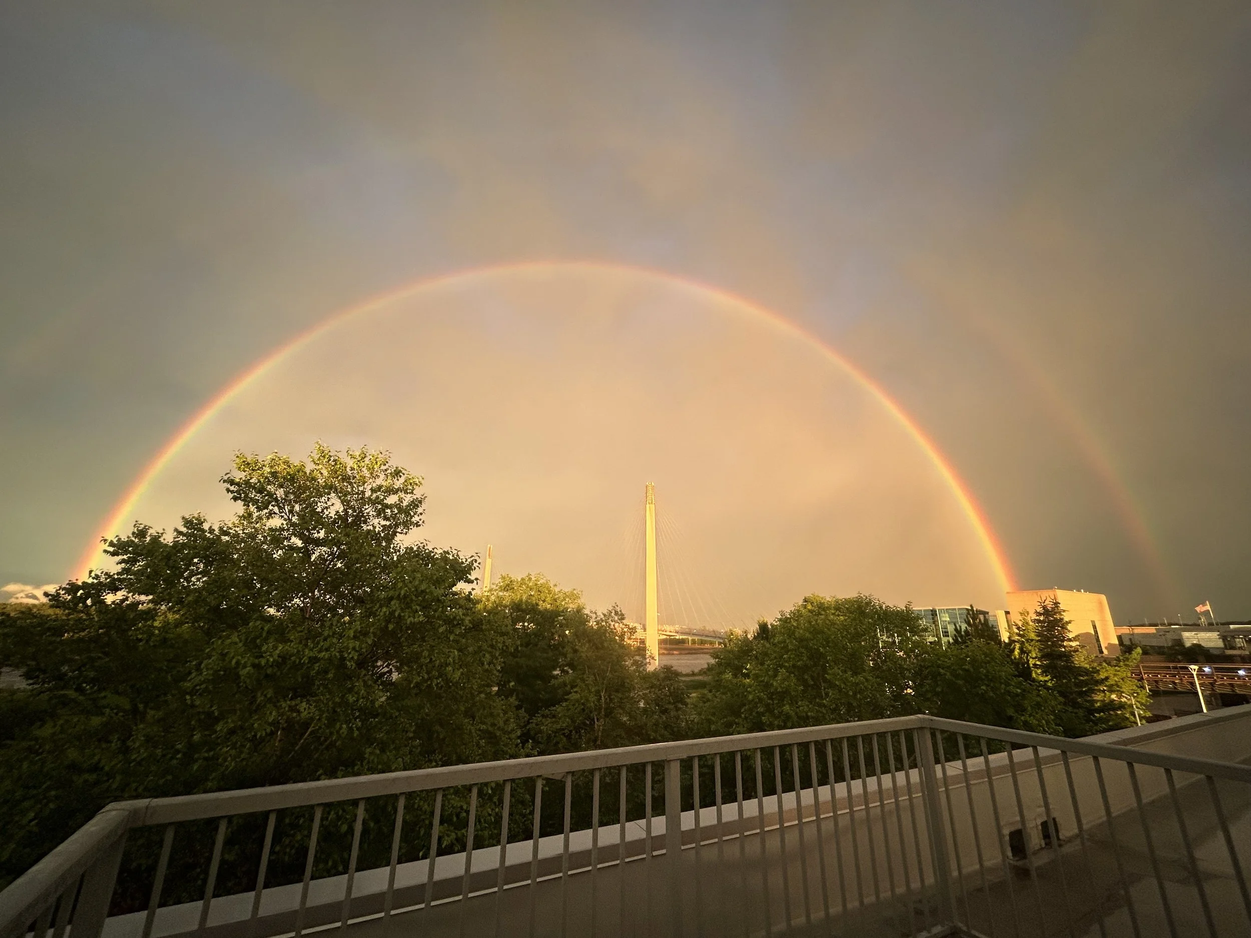 Rainbow arching over trees and buildings during a sunset, with a cloudy sky and a cityscape in the background, viewed from a balcony with a white railing.
