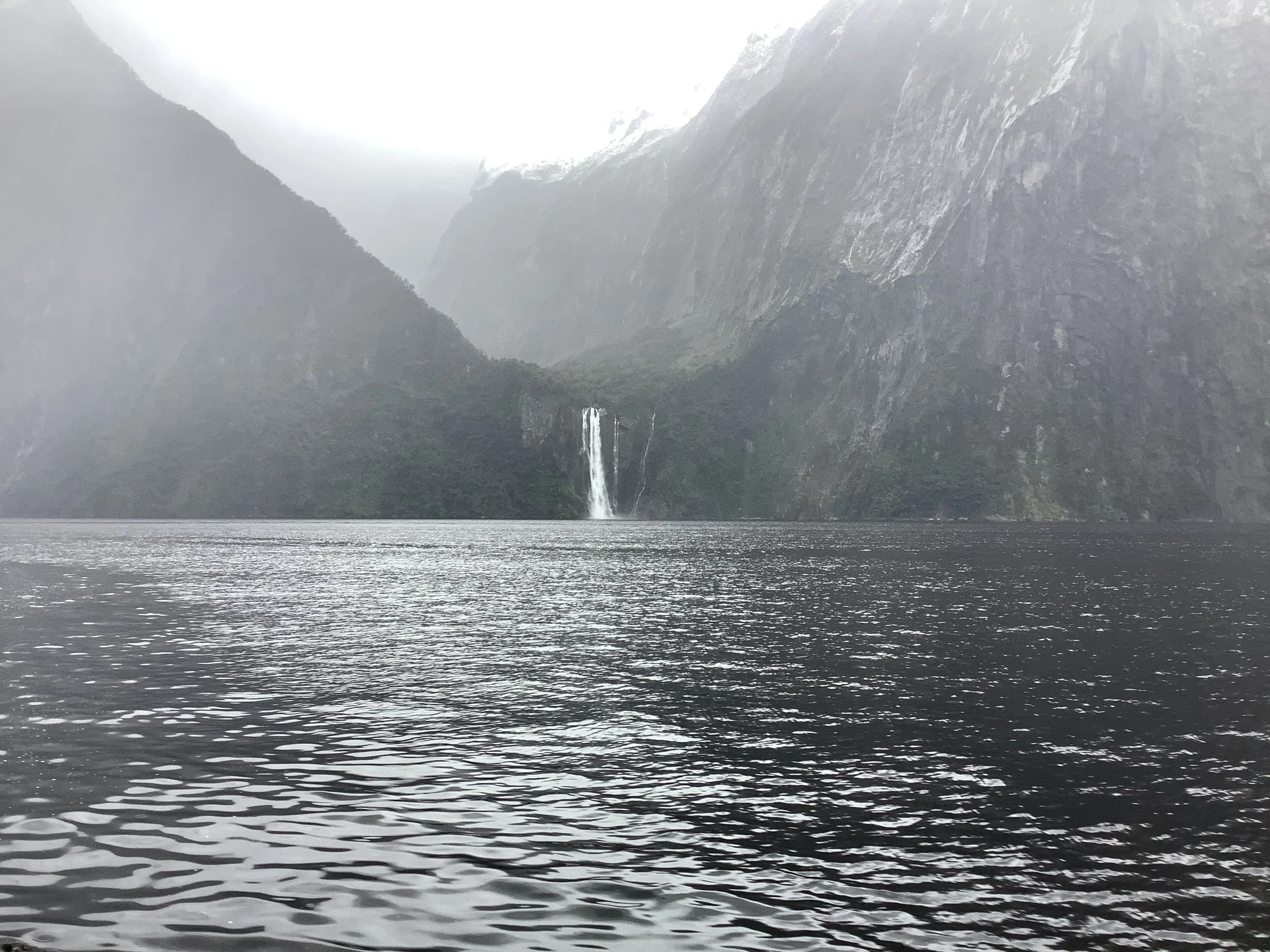 A body of water in a valley with steep, misty mountains on either side and a waterfall in the distance.