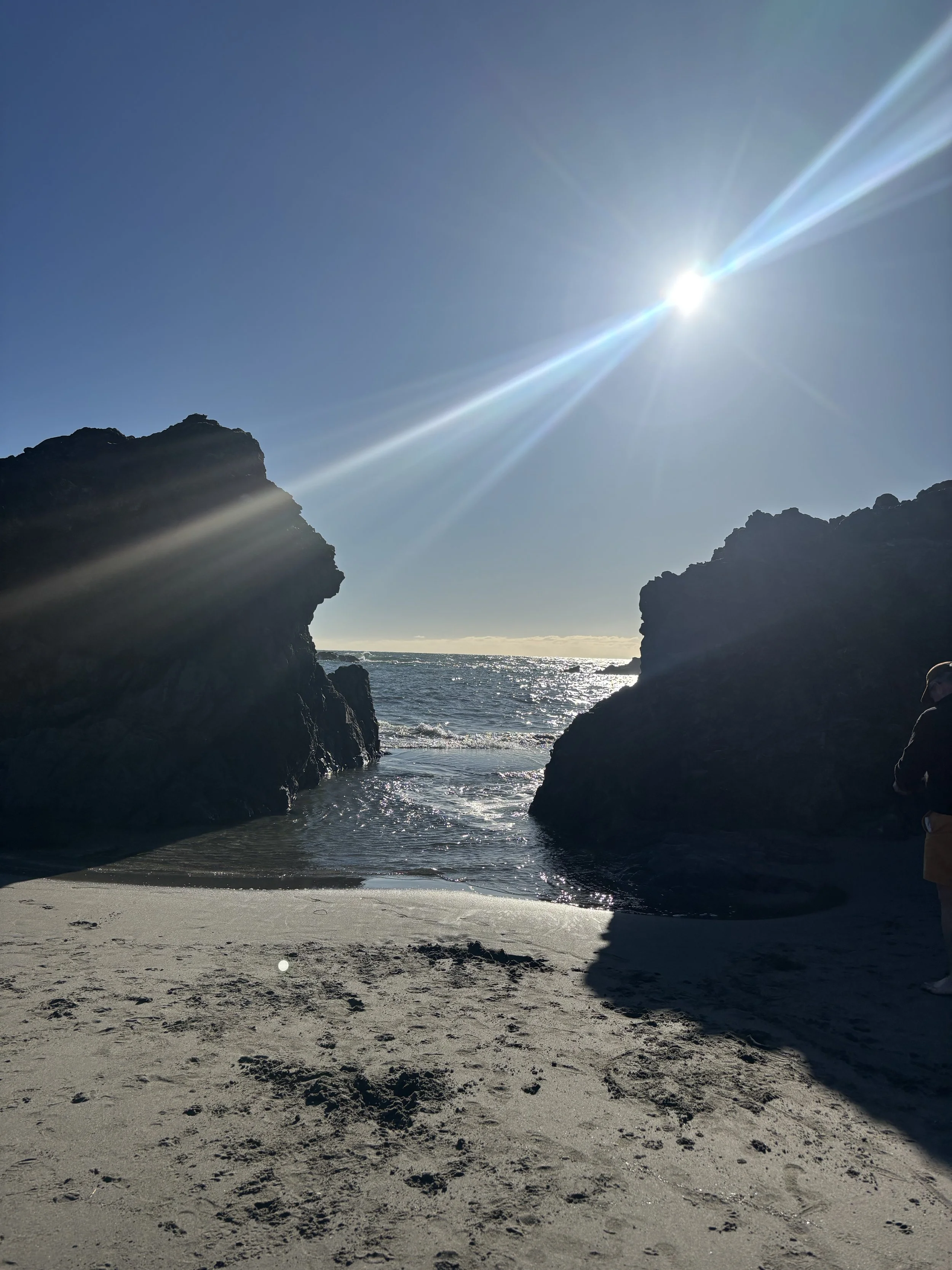 A beach scene with large rock formations framing the ocean and the sun shining brightly in a clear blue sky.