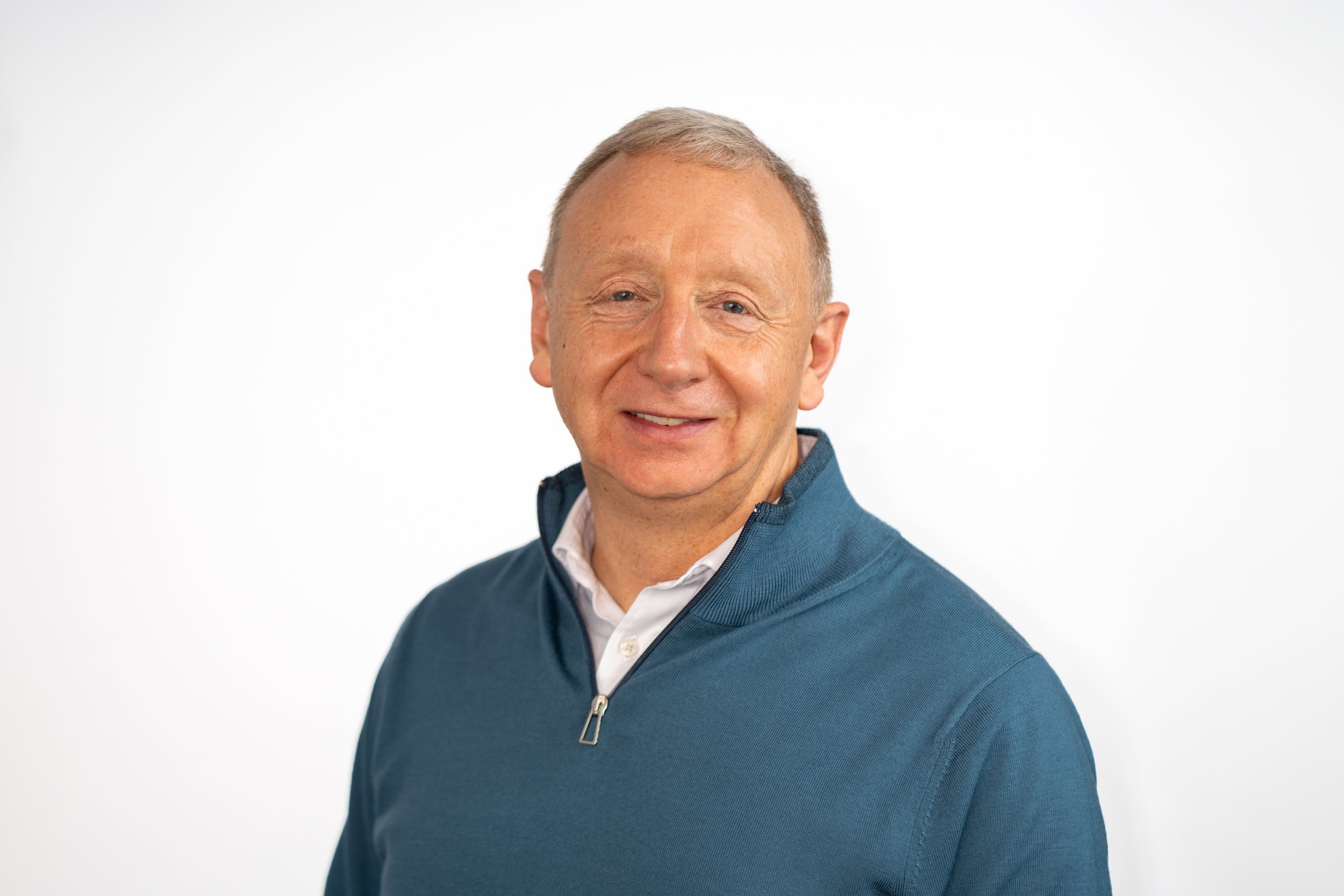 Headshot of a smiling middle-aged man with short gray hair, wearing a white collared shirt underneath a blue quarter-zip sweater, against a plain white background.