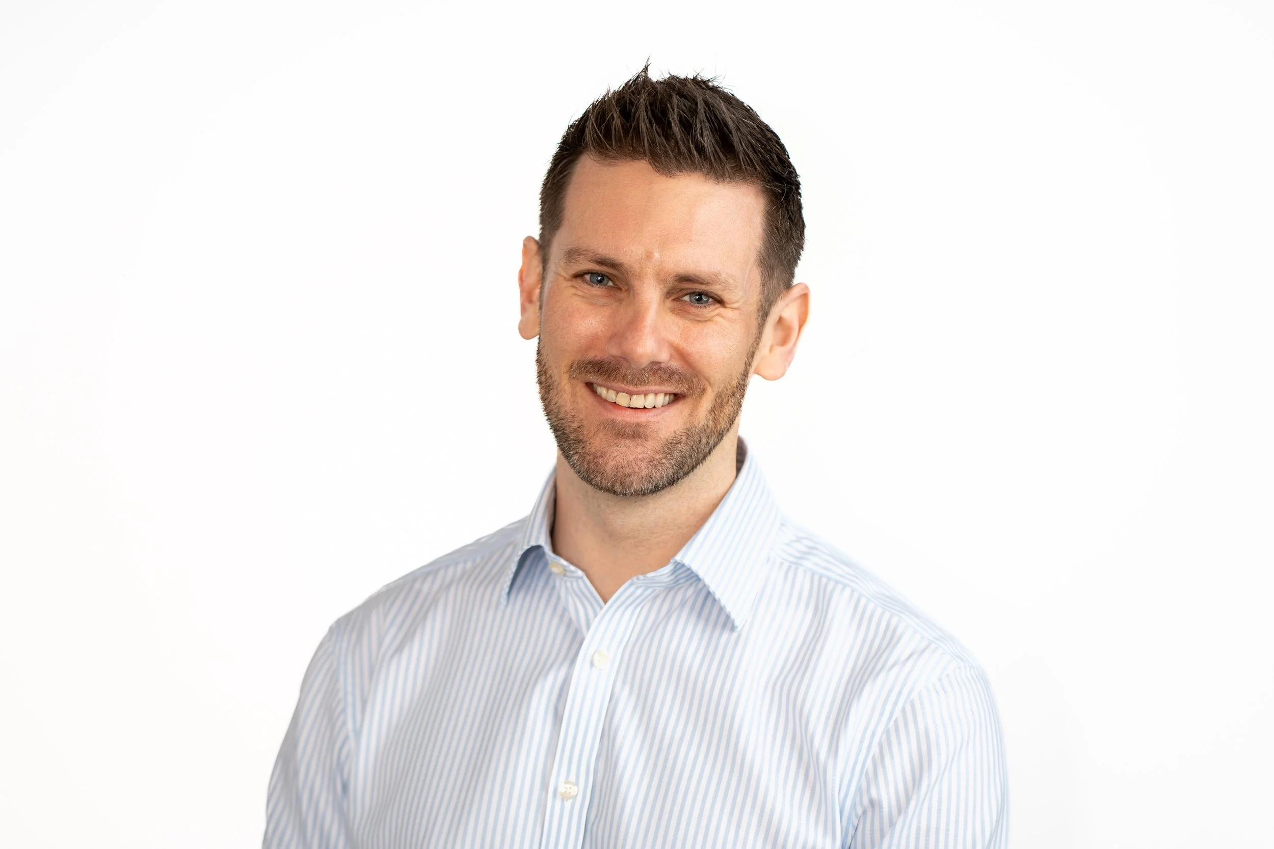 Smiling man with short dark hair, beard, wearing a light blue striped button-up shirt, against a white background.