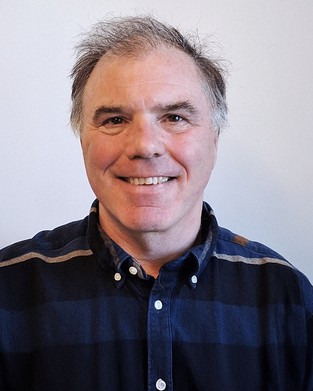 Headshot of a smiling man with short, graying hair, wearing a dark blue button-up shirt with a beige stripe on the shoulders, standing against a plain white background.