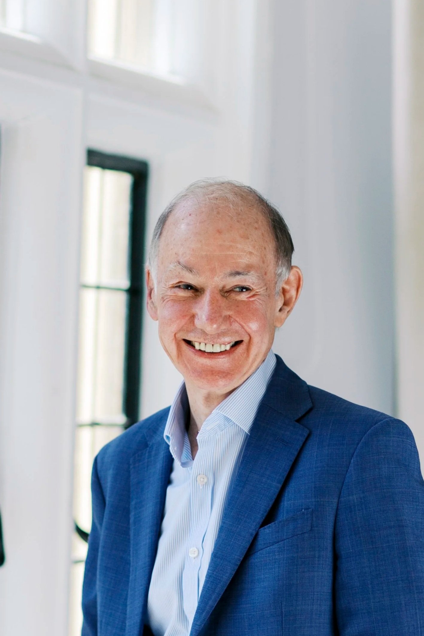 A smiling older man in a blue suit and white shirt, standing indoors near windows with white walls.