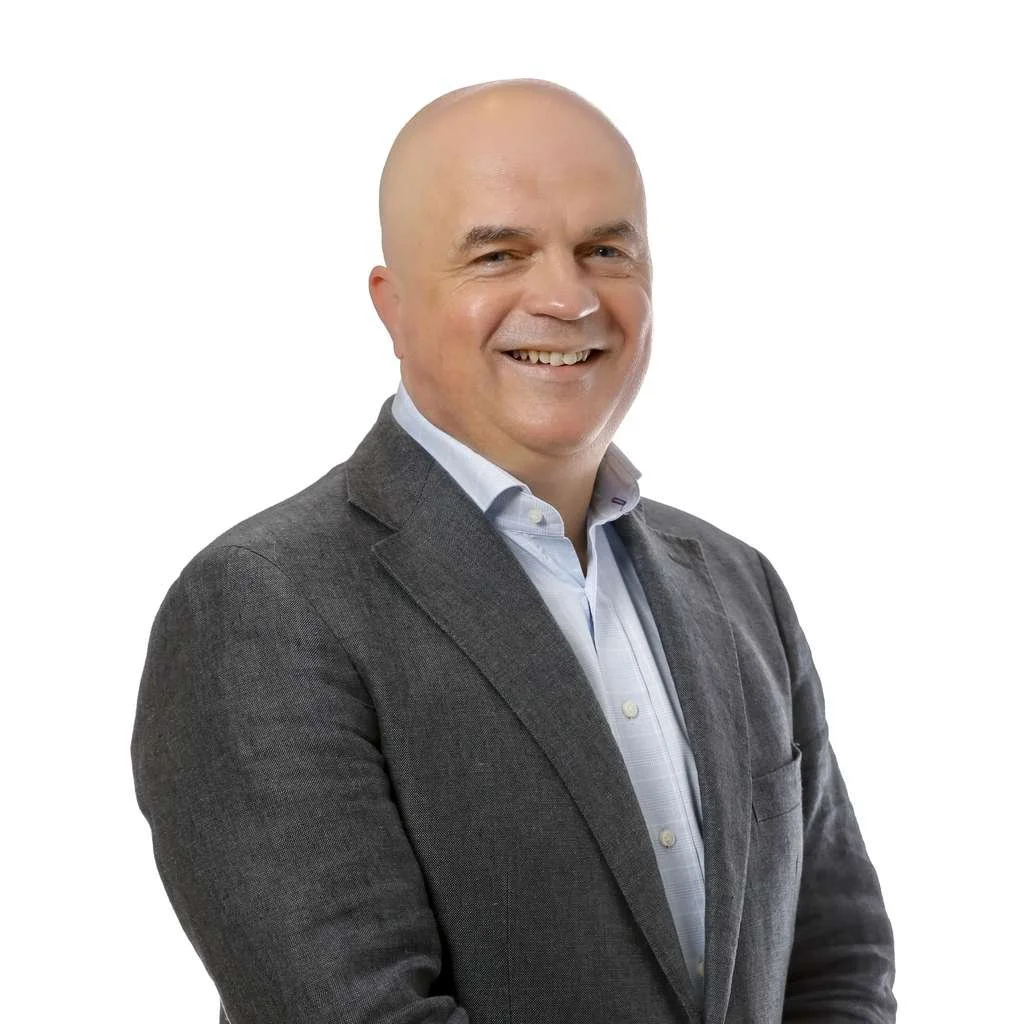 A smiling middle-aged man with a bald head, wearing a dark gray blazer over a light blue dress shirt, standing against a white background.