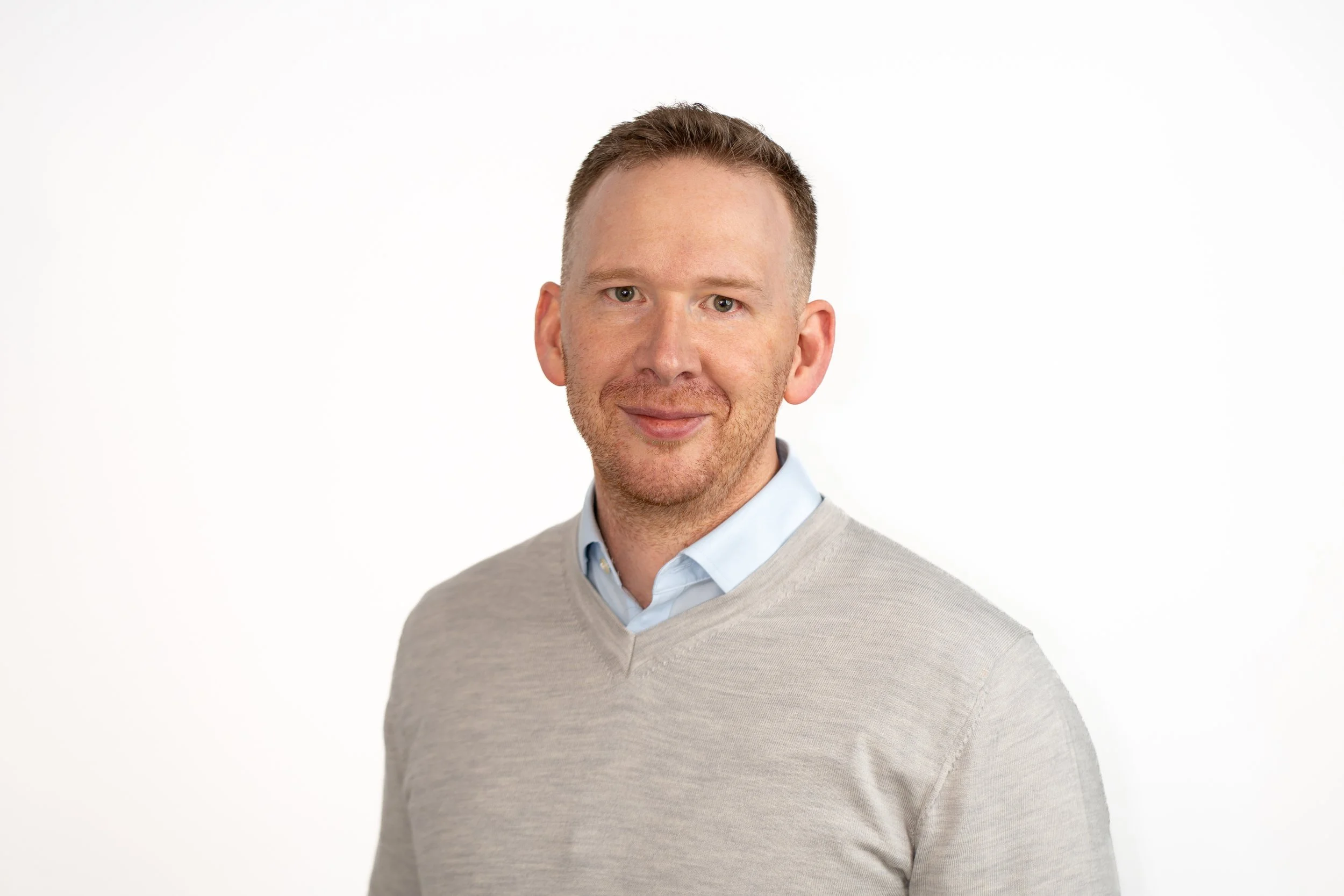 A man with short brown hair, light skin, and a light beard, wearing a light blue collared shirt and a gray sweater, smiling slightly against a plain white background.