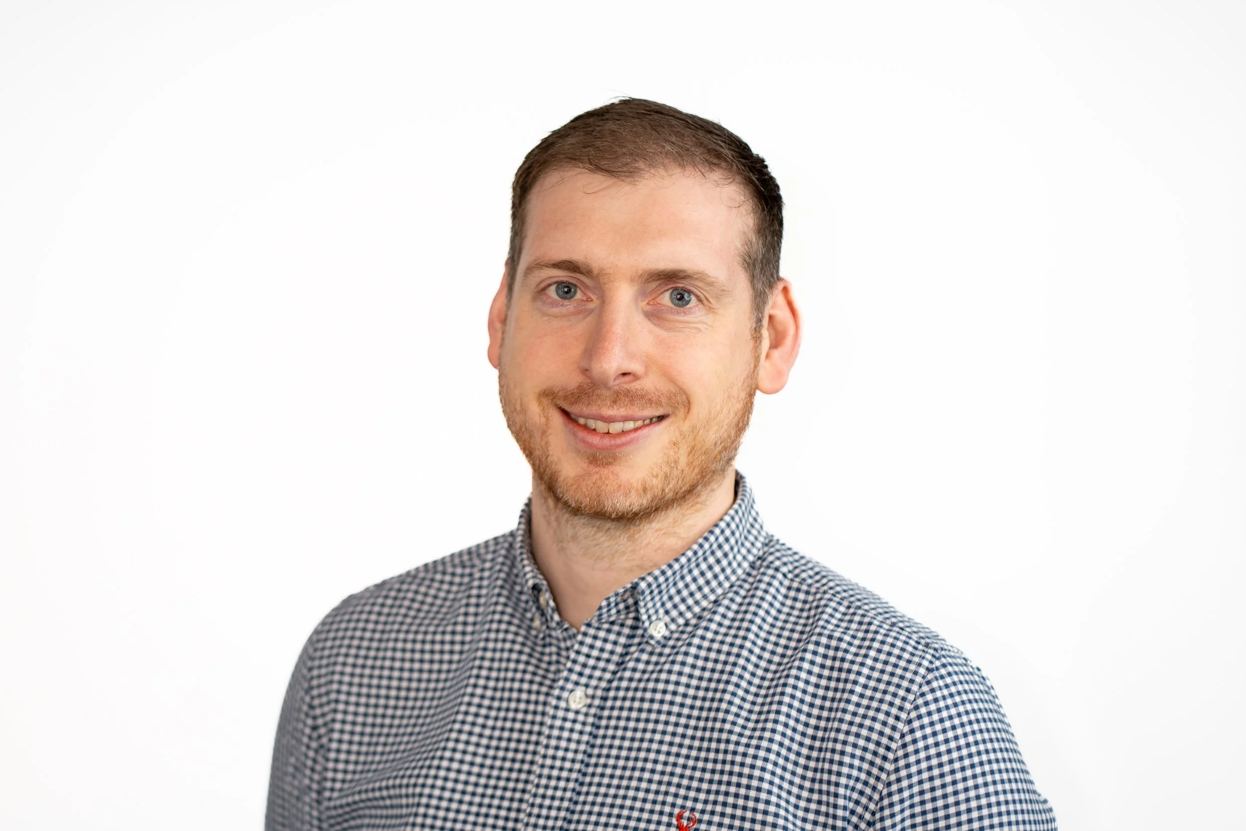Portrait of a smiling man with short brown hair, blue eyes, a light beard, wearing a checkered button-up shirt, against a plain white background.