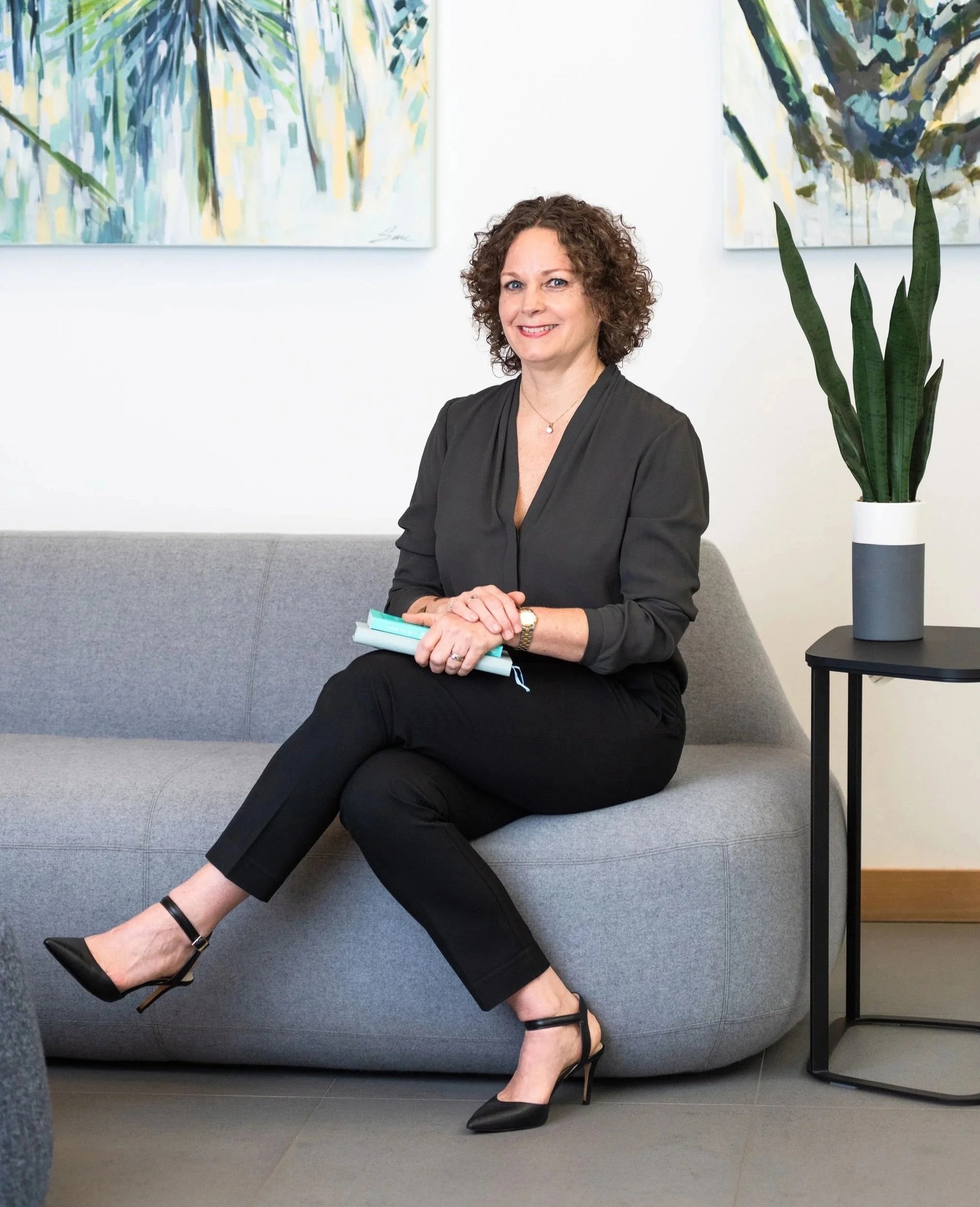 Kate Hanley wearing a black blazer and pants, sitting on a gray sofa, holding some papers, with a large green leafy plant in a white pot on a side table next to her and abstract paintings with green and yellow hues on the white wall behind her.