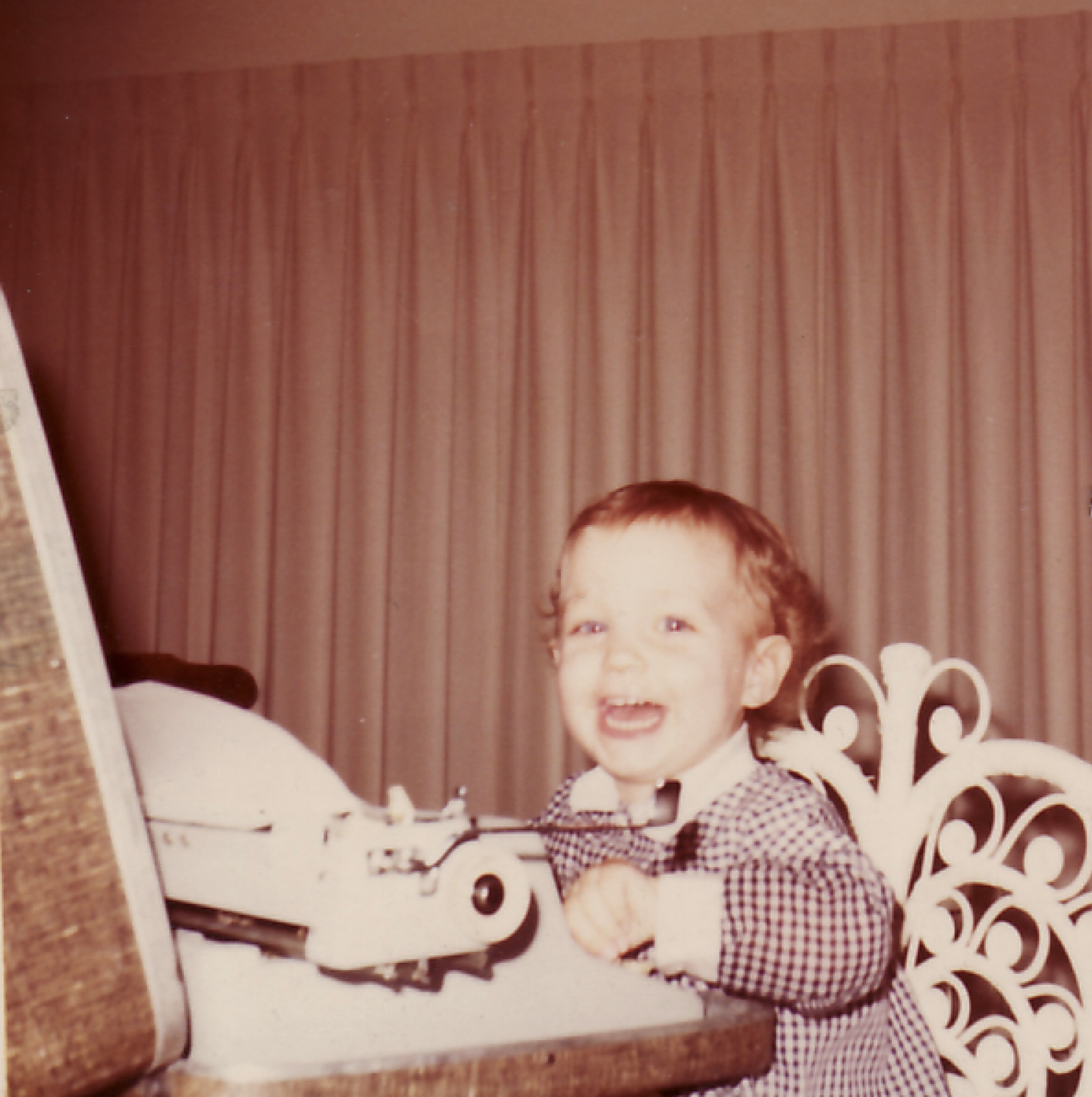 Kate Hanley as a young child with short curly hair and a checkered dress sitting in a white ornate metal bed, with a smile and an open mouth, in a room with brown striped curtains.