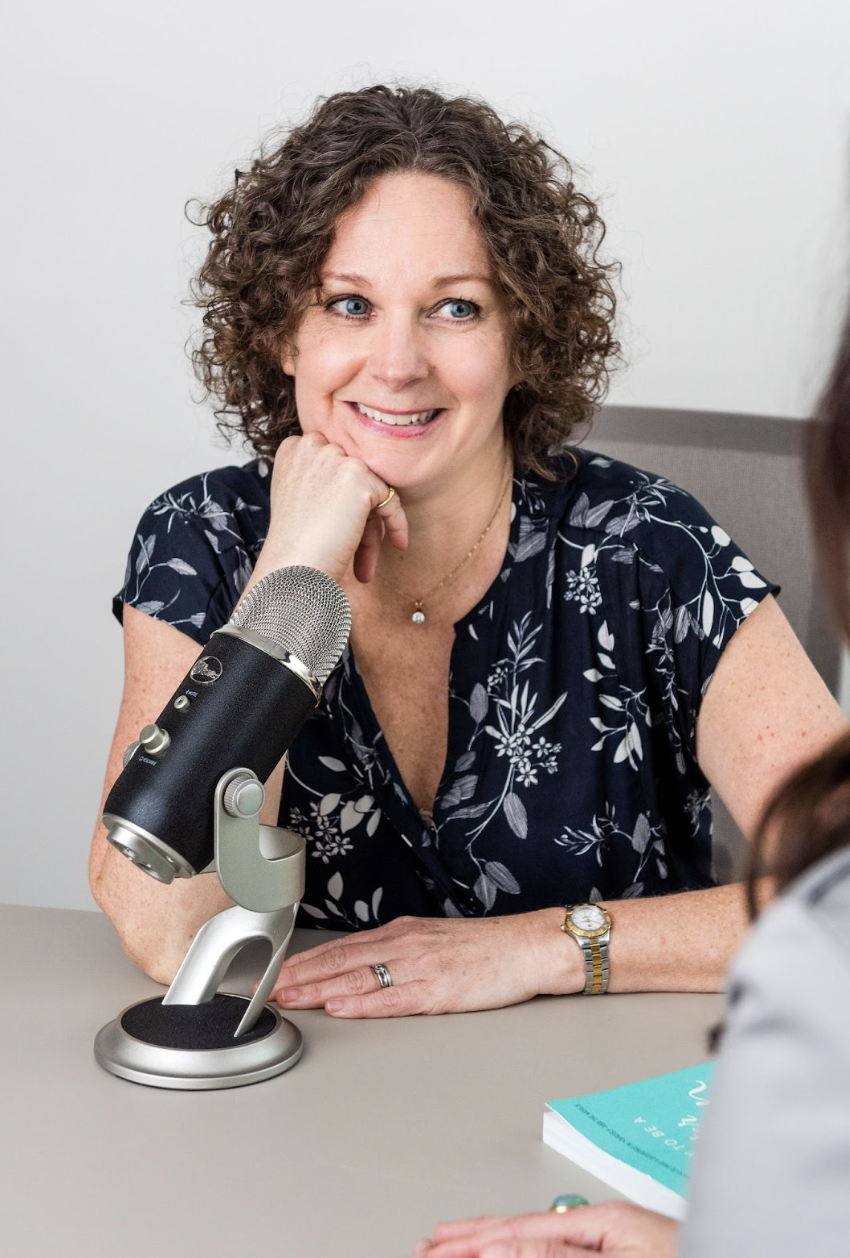 Kate Hanley with curly brown hair, wearing a dark floral shirt, sitting at a table with a microphone in front of her, smiling with her chin resting on her hand.