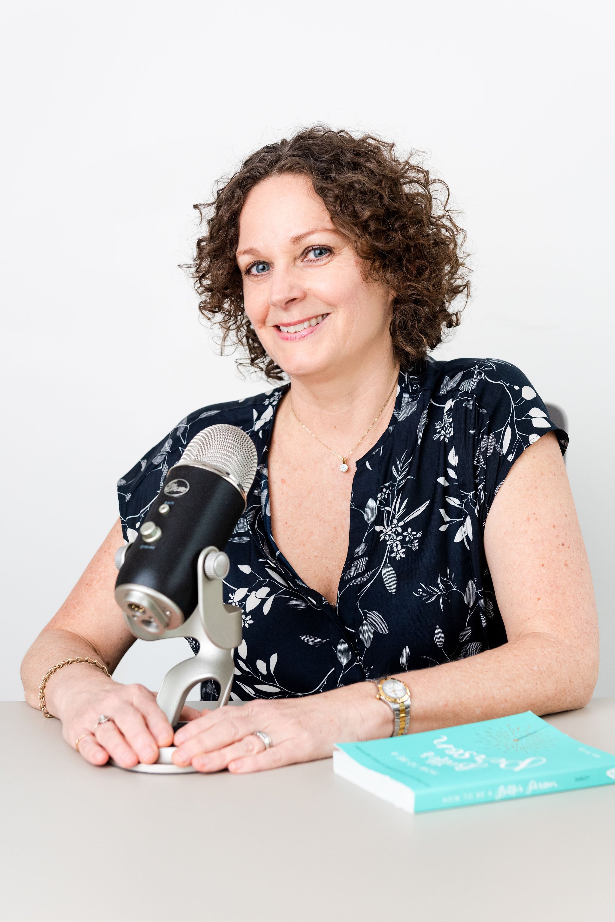 Kate Hanley wearing a navy dress with white floral pattern, sitting at a table with a book titled "How To Be a Better Person" in front of her, smiling, with a professional microphone on a stand in front of her, and her hands resting on the table.