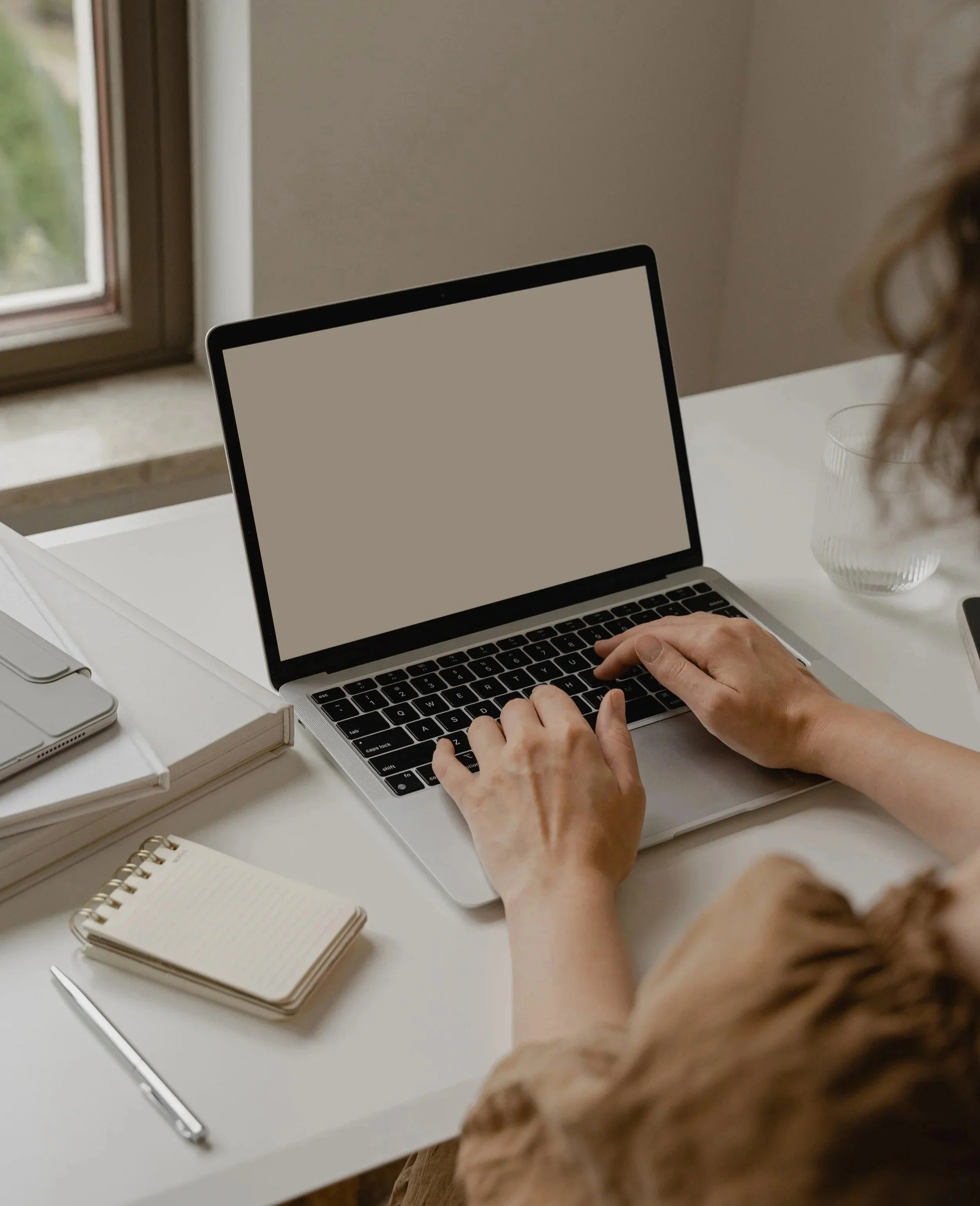 Person with curly hair using a laptop at a white desk near a window with greenery outside.