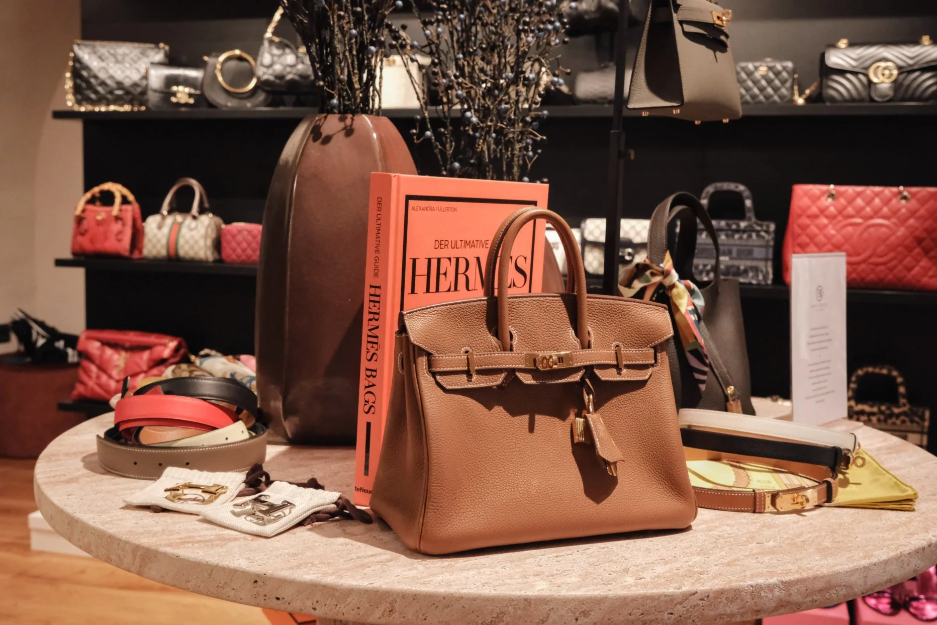 Auf einem runden Tisch befinden sich mehrere Designer-Taschen, darunter eine beige Birkin-Tasche und eine schwarze Kelly-Tasche, daneben ein pinkes Buch mit dem Titel 'Der Ultimative Hermès Bags Guide'. Im Hintergrund sind Regale mit weiteren Handtaschen und kleinen Ledertaschen zu sehen.