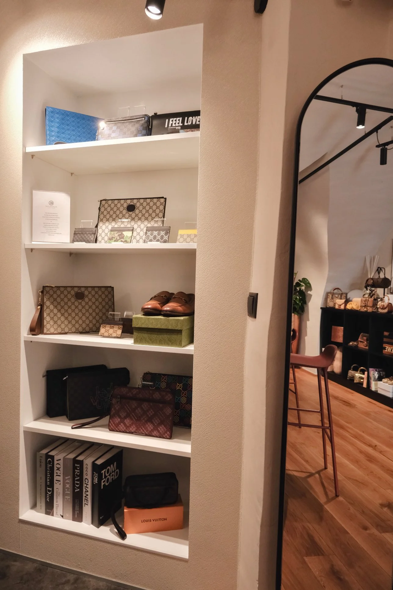 Gerahmte Taschen und Schuhe auf weißen Regalen in einem Modegeschäft, Blick in einen Sitzbereich mit Stühlen und Regal mit Taschen im Hintergrund.