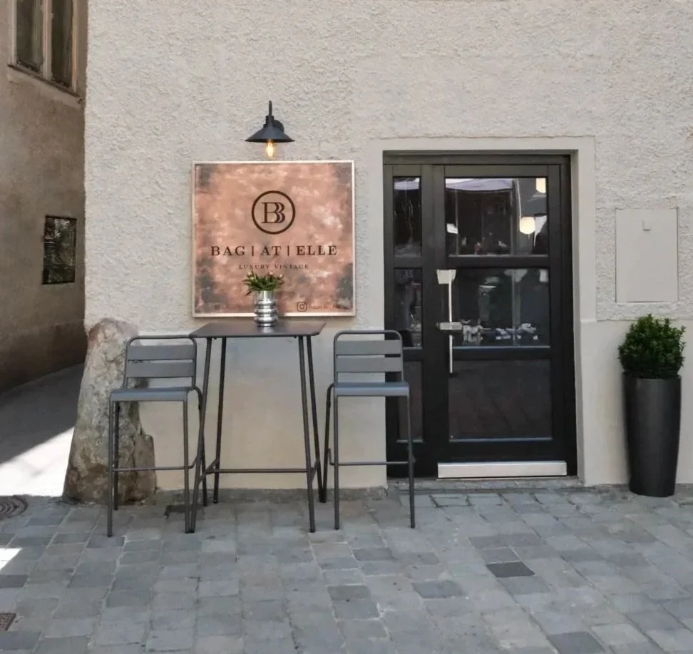 Außenansicht eines Eingangsbereichs mit einer schwarzen Tür, einem Blumenpotted, einem hohen Tisch mit zwei Hockern, einer Wandlampe und einem Firmenlogo an der Wand, das 'BAGATELLE' mit der Aufschrift 'Luxury Vinage' zeigt.