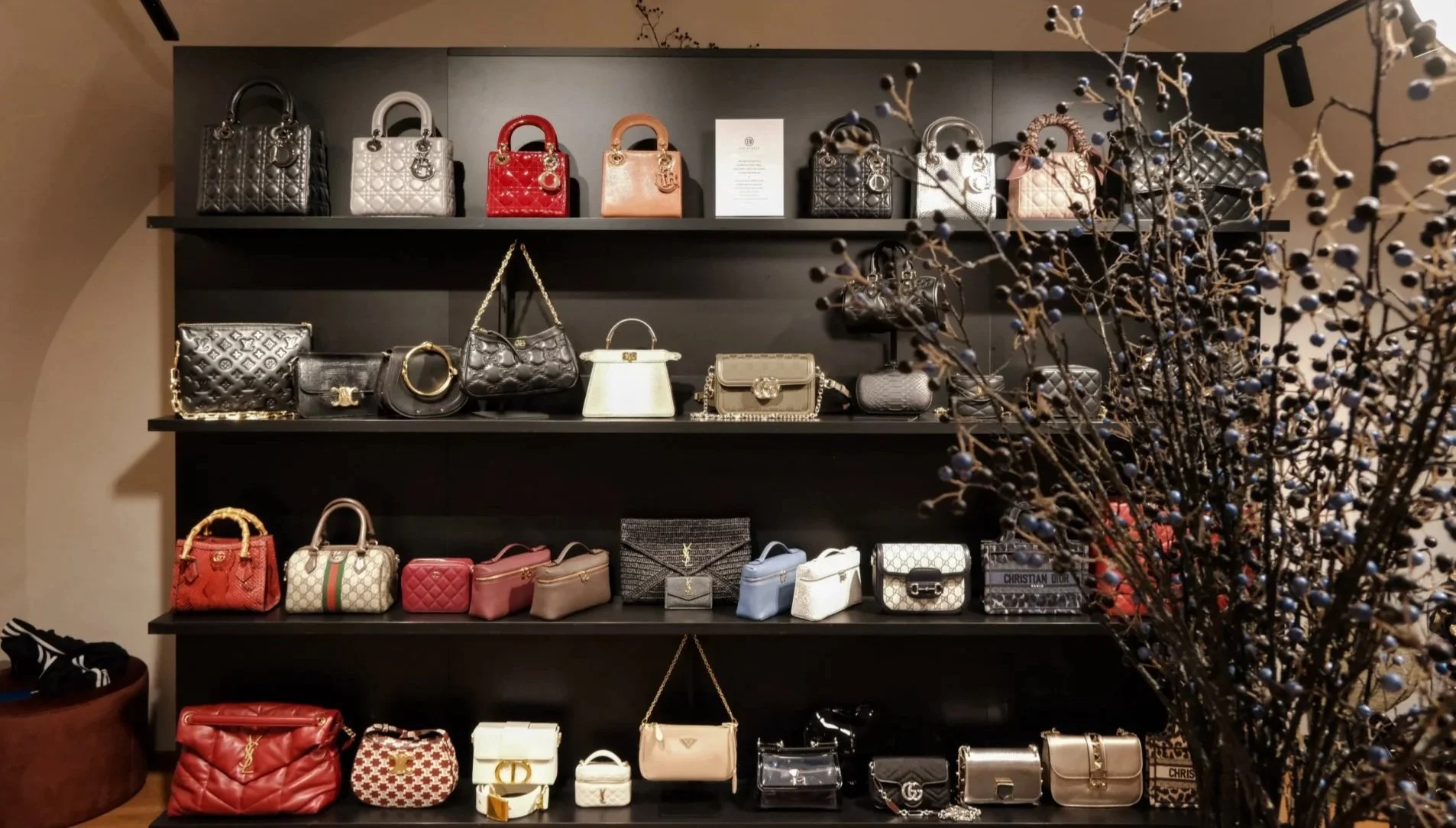 Regal mit einer Vielzahl von Designer-Taschen, hauptsächlich in Schwarz, Weiß, Rot, Beige und Grau, auf schwarzen Regalbfüßen. Ein Blumenstrauß mit schwarzen Beeren ist im Vordergrund sichtbar.