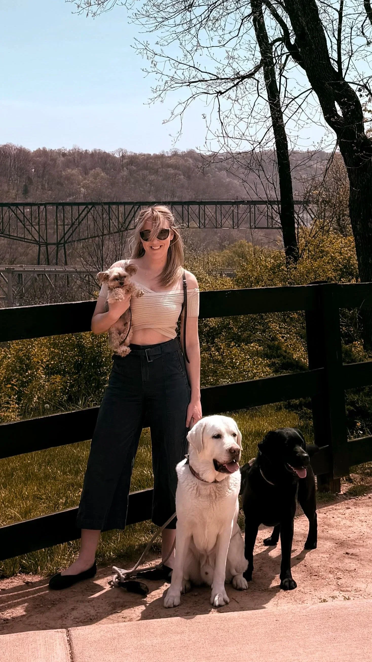 A woman with blonde hair wearing sunglasses, a beige off-the-shoulder top, and black pants, holding a small dog, standing outdoors next to two large dogs, one white and one black, near a black fence with trees and hills in the background.