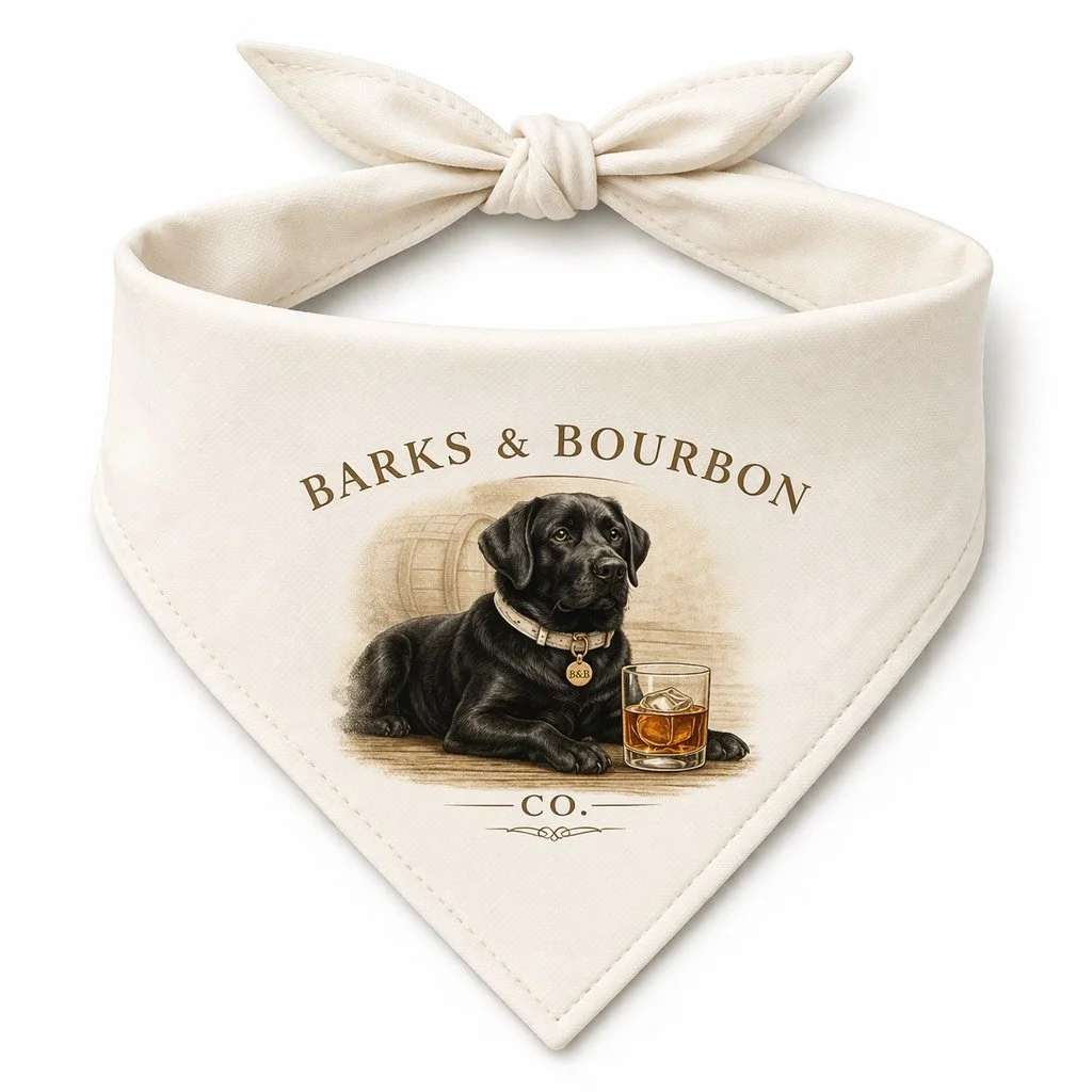 A cream-colored bandana with a printed design featuring a black Labrador Retriever dog wearing a collar with a tag, sitting next to a glass of bourbon with ice, and a background with a barrel. The text reads "Barks & Bourbon Co."