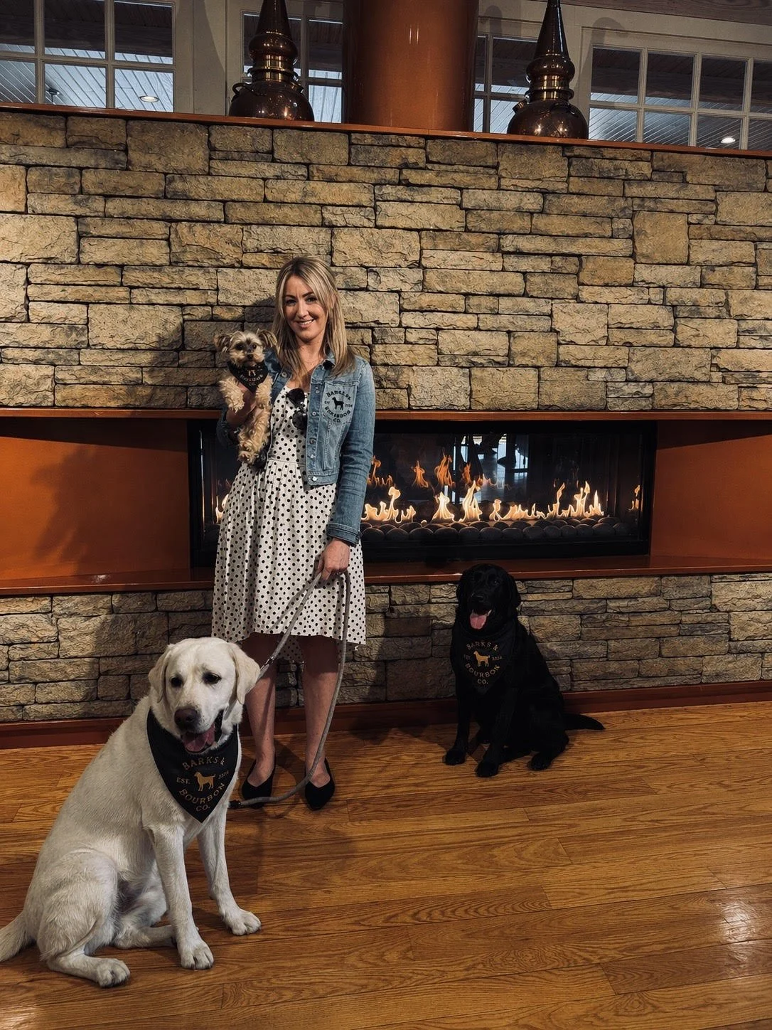 A woman in a polka dot dress and denim jacket holds a small dog while standing next to a large fire feature in a room with wooden floors and a stone and wood fireplace. Two other dogs, one yellow Labrador and one black, sit on the floor near her, all wearing matching scarves with a logo.