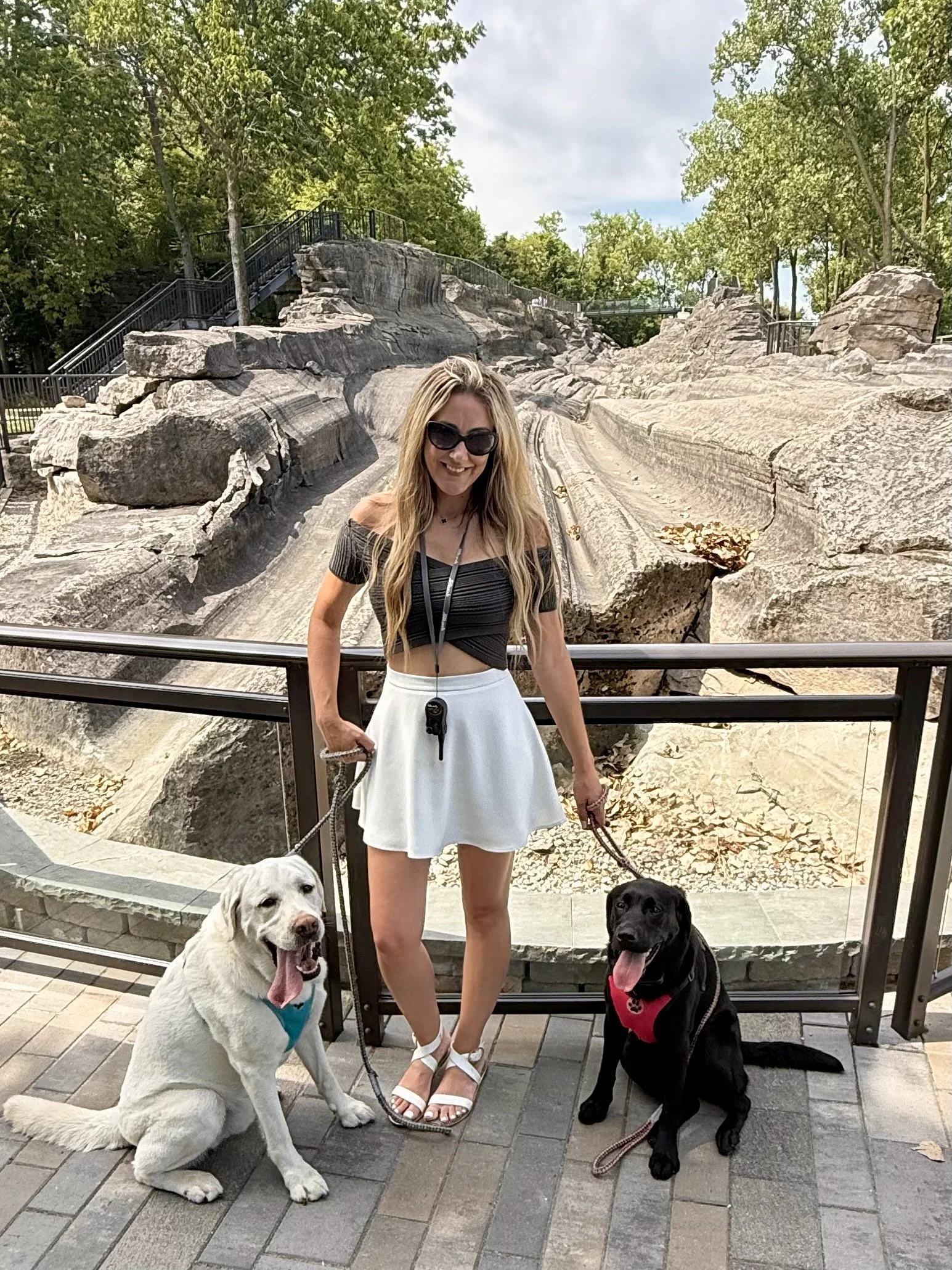 A woman with long blonde hair and sunglasses standing outdoors on a stone pathway, holding leashes of two dogs, one yellow Labrador Retriever and one black Labrador Retriever. Behind her are large rocks and trees.