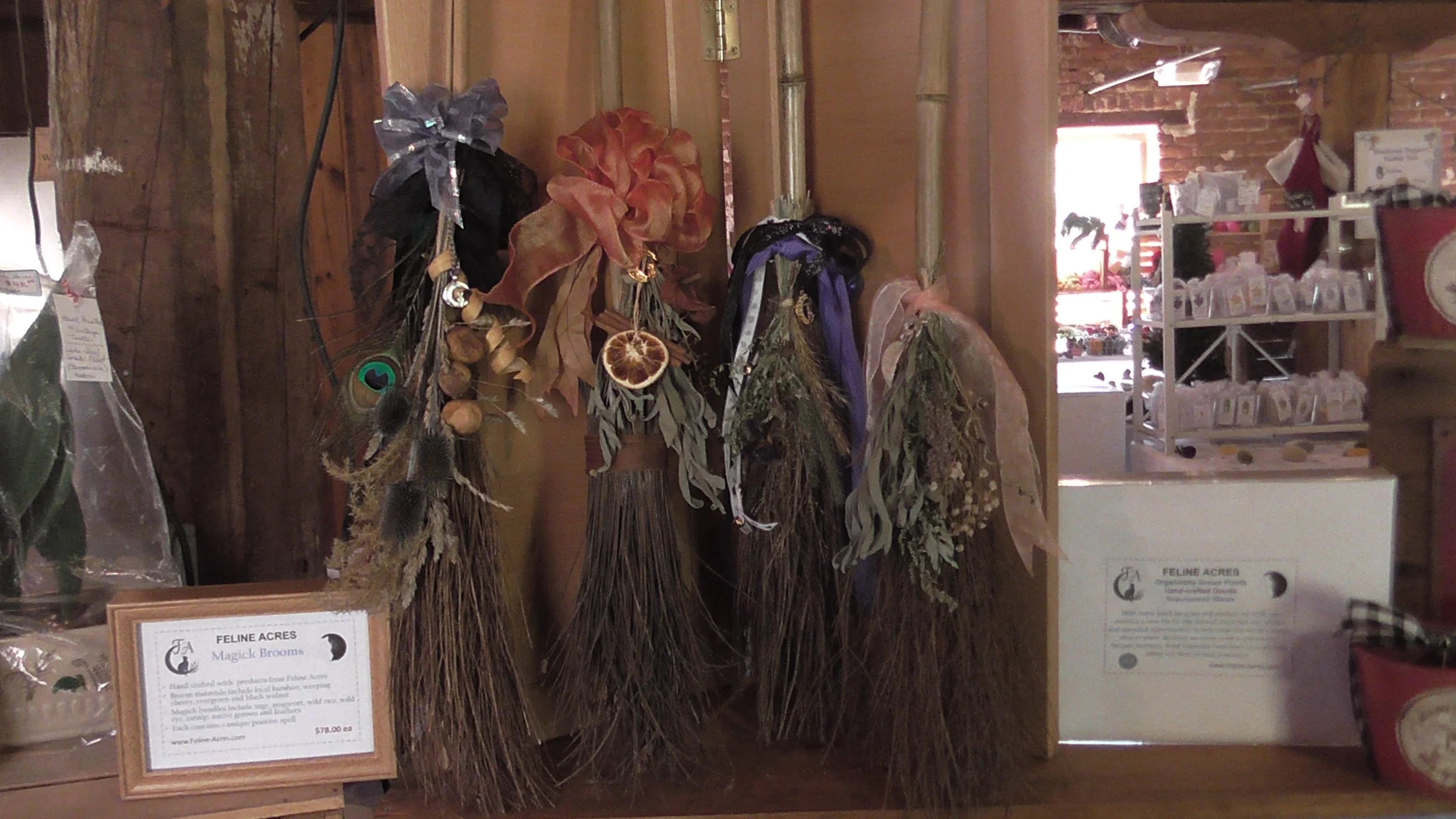 Display of decorated magick witches broomsticks with dried flowers, leaves, and natural elements in a rustic store setting. There is a sign mentioning 'Magick Brooms' and a price of $78.