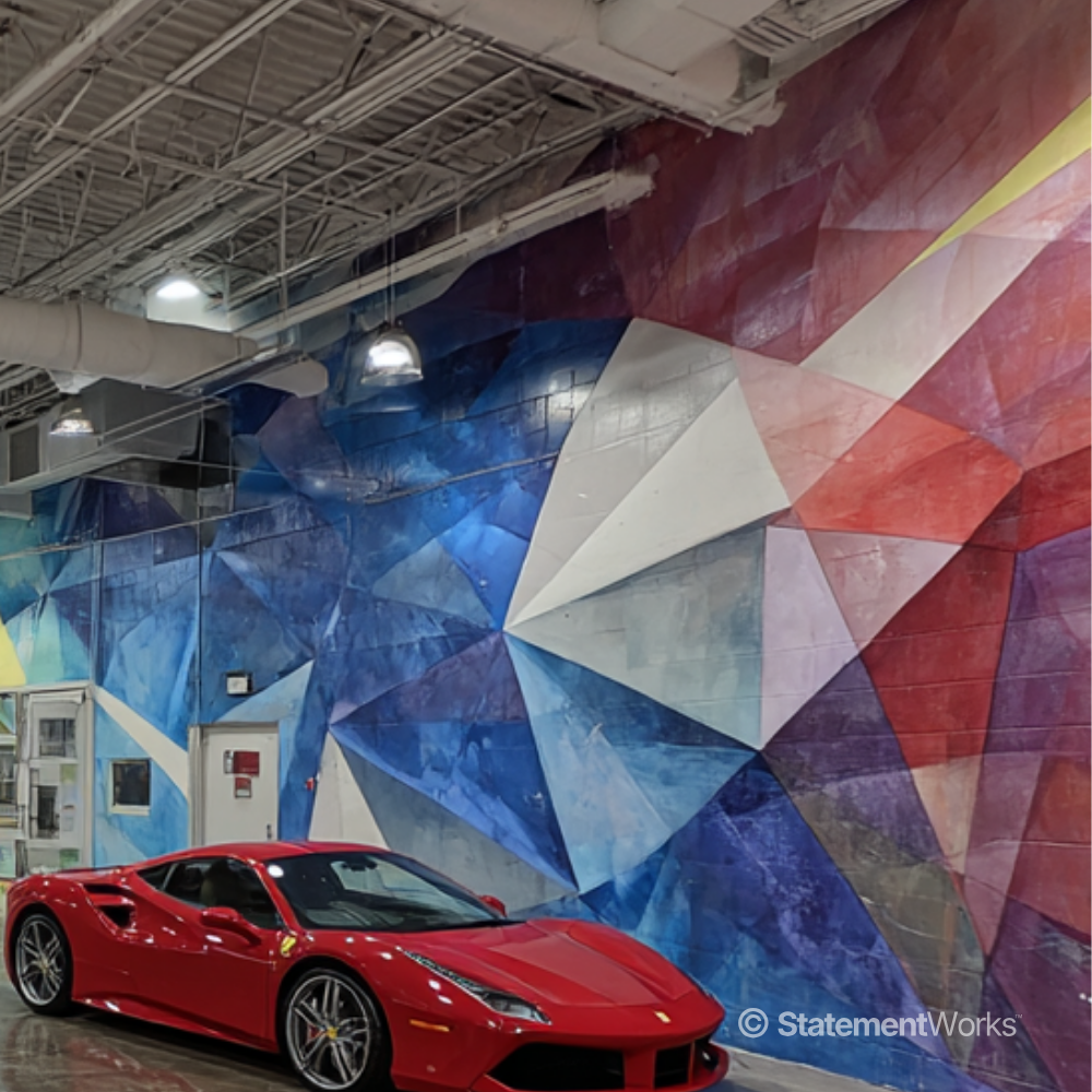 A red Ferrari sports car parked inside a garage with a colorful, geometric mural on the wall featuring shades of blue, white, red, purple, and yellow, and visible industrial ceiling lighting and ductwork.
