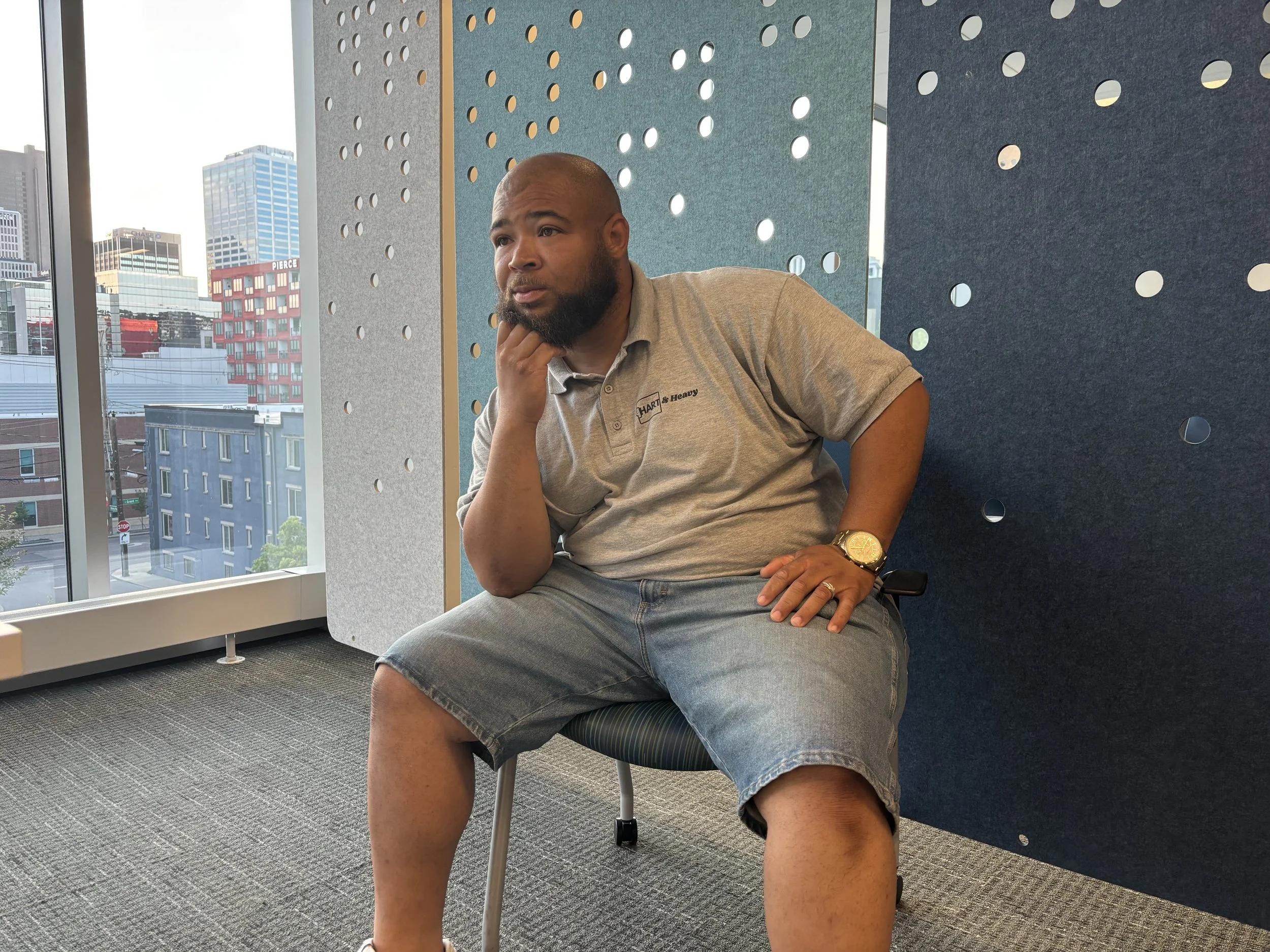 A man sitting on a chair in an office, looking thoughtful, with a cityscape visible through large windows behind him and a blue perforated wall.