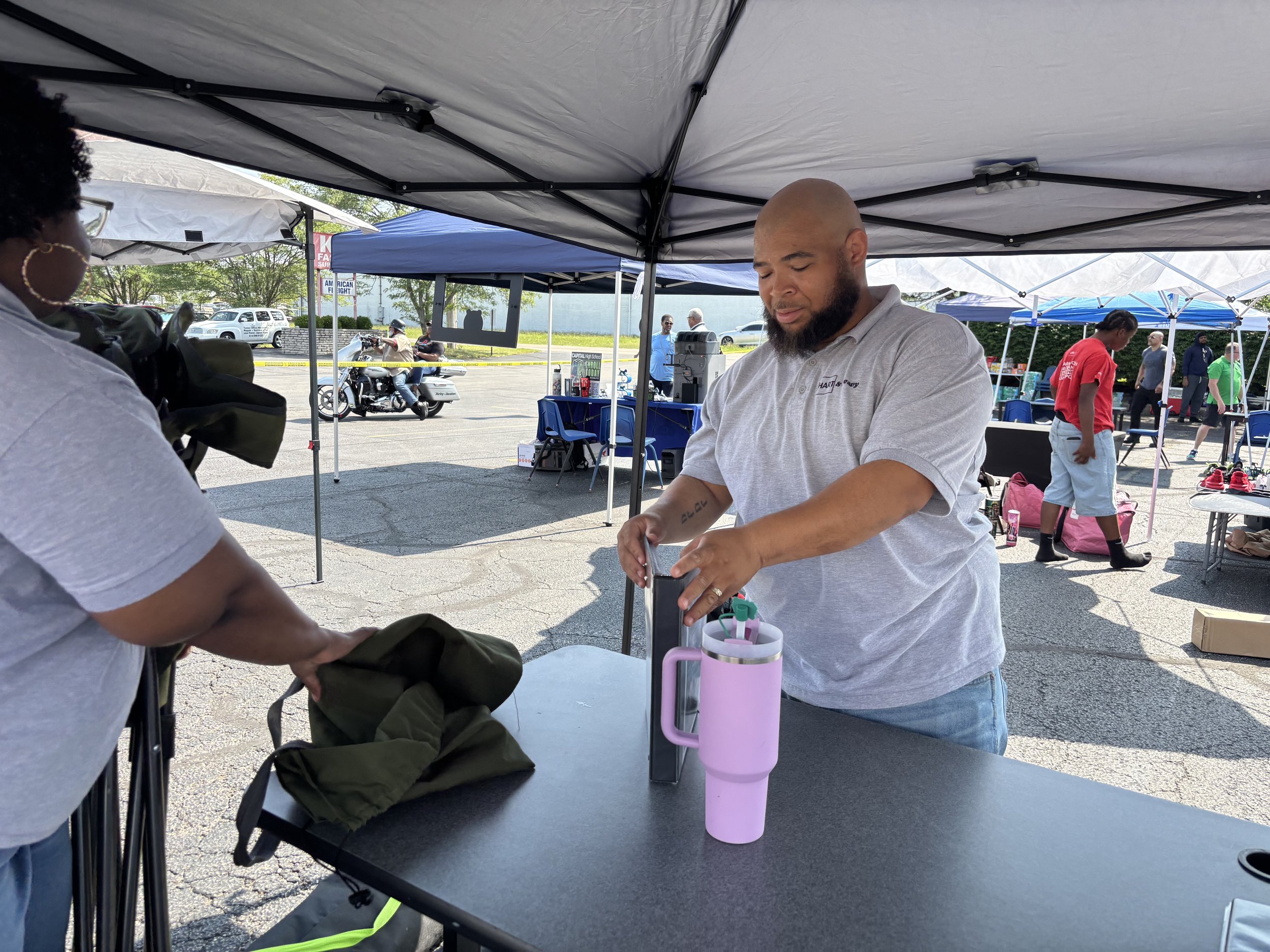 A man with a beard and a bald head wearing a gray polo shirt is standing under a canopy at an outdoor event, working on something on the table. There are other tents, people, and trees in the background.