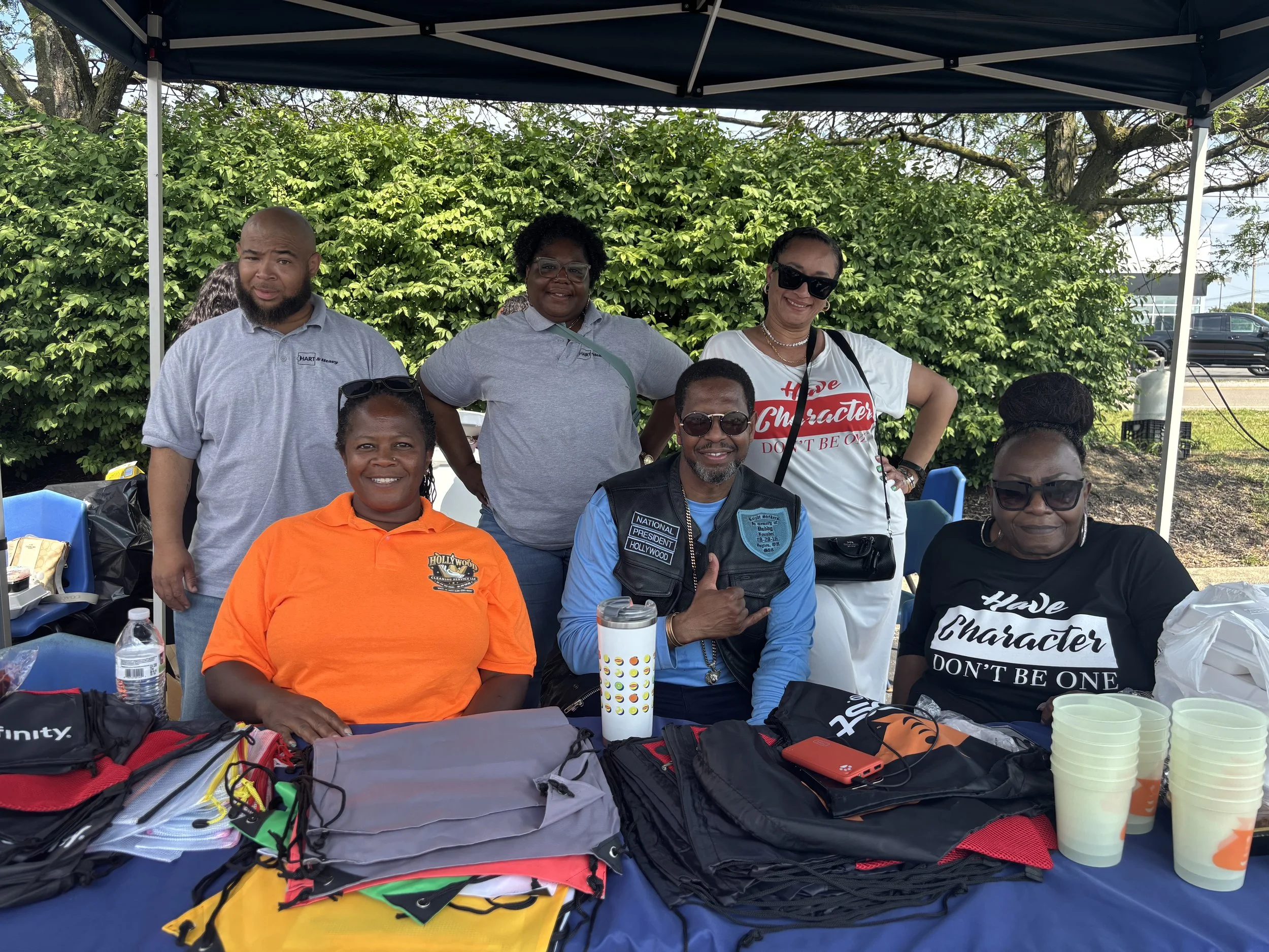 Group of people gathered under a blue canopy outdoors, smiling at the camera with a table of items including water bottles, cups, and clothing in front of them.