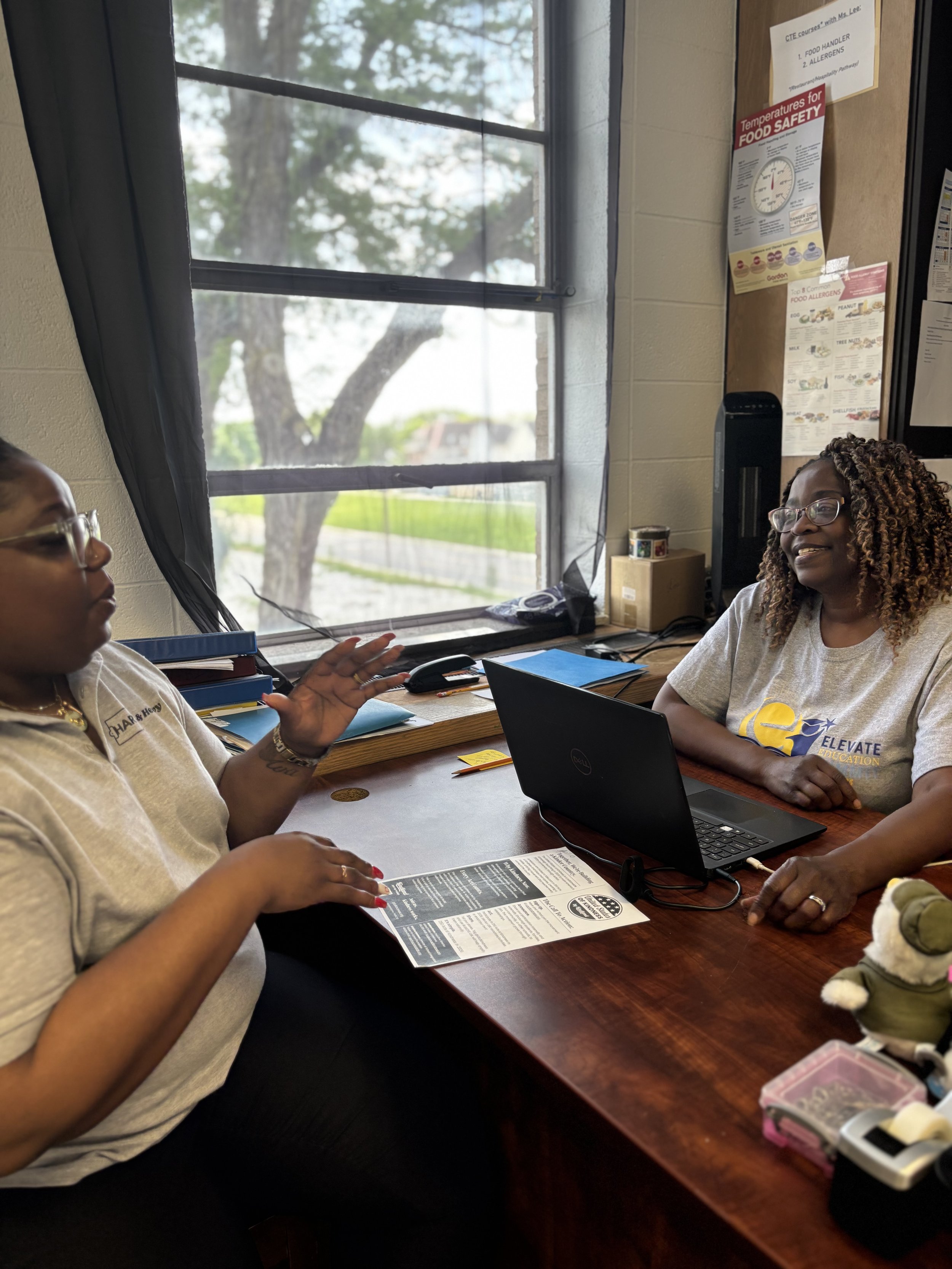 Two women sitting at a wooden table having a conversation in a room with large window showing trees outside. One woman has glasses, curly hair, and is wearing a T-shirt with a logo. The other woman is wearing glasses and a beige shirt. The table has a laptop, papers, and office supplies.