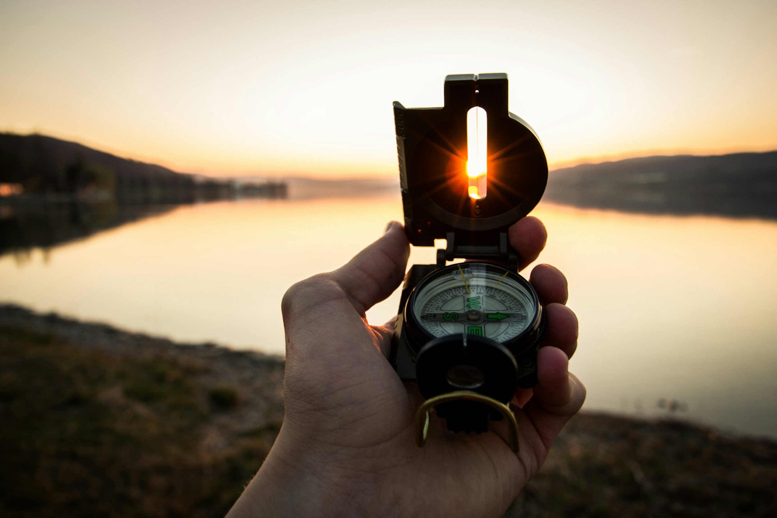 A person holds a compass while taking a photograph at sunset over a lake with distant hills, with the sunlight shining through the compass.