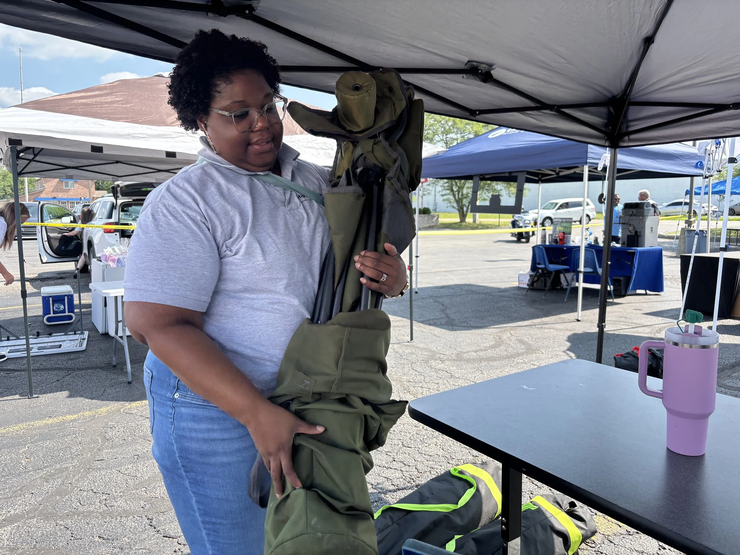 A woman with glasses and curly hair setting up camping chairs at an outdoor event under a large canopy. There are tables, chairs, and other people in the background.