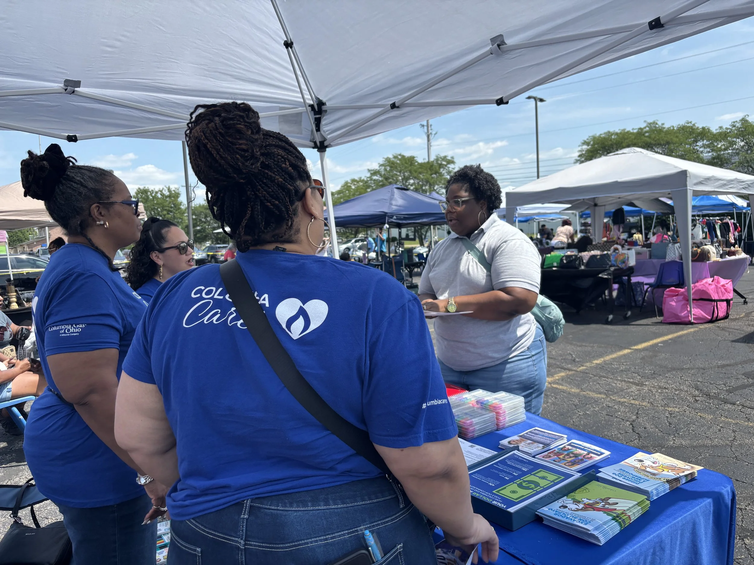 Group of women at an outdoor booth, talking and looking at pamphlets with tents and other booths in the background.