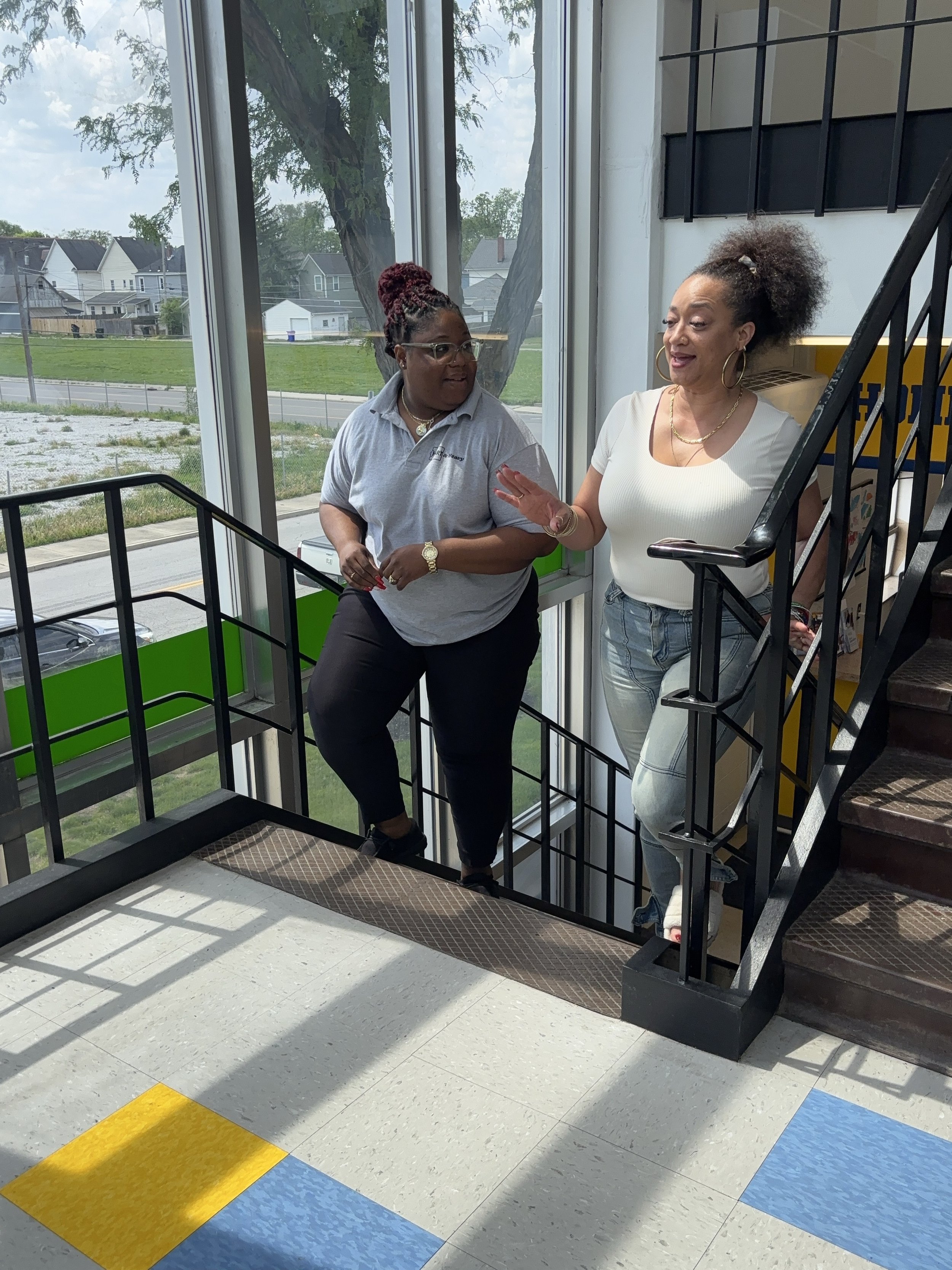 Two women having a conversation at the top of a staircase inside a building, with large windows in the background showing trees and houses outside.