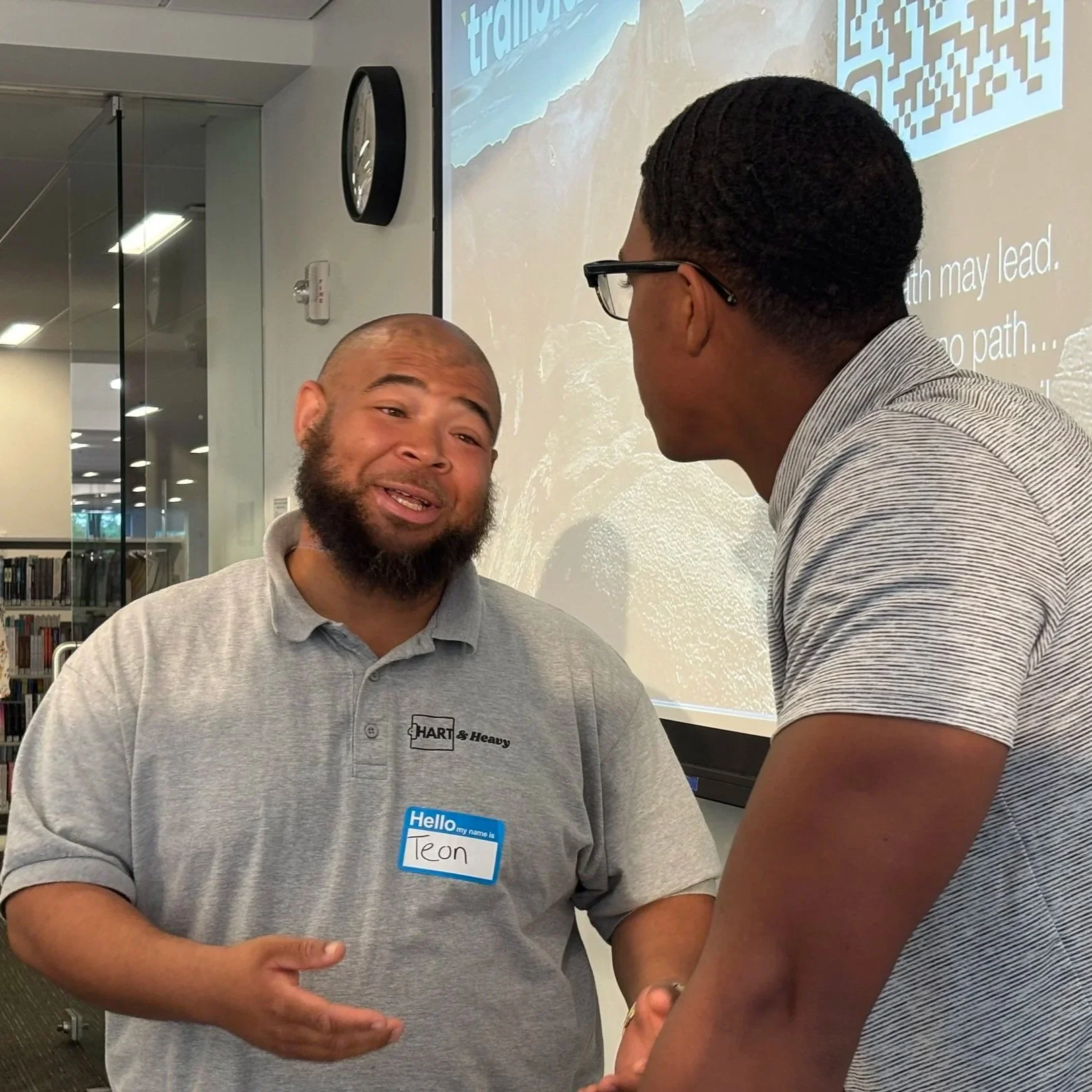 Two men are engaged in a conversation in an indoor setting. The man on the left wearing a gray polo shirt is smiling and gesturing with his hands. The man on the right, wearing glasses and a striped shirt, is facing him. In the background, there's a white screen with text and a QR code, a black wall clock, and books on shelves.