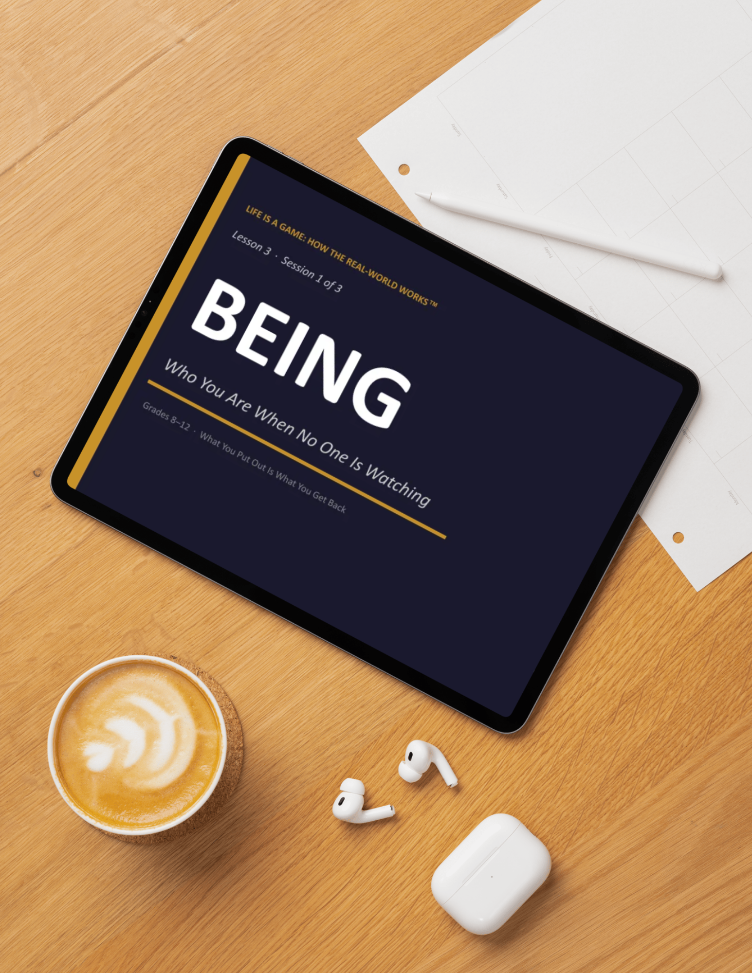 A flat lay of a wooden desk with a tablet showing a slide titled 'BEING' and text about life as a game, a white pen, a sheet of paper with grid lines, a cup of latte with latte art, white wireless earbuds, and a white wireless earbud case.