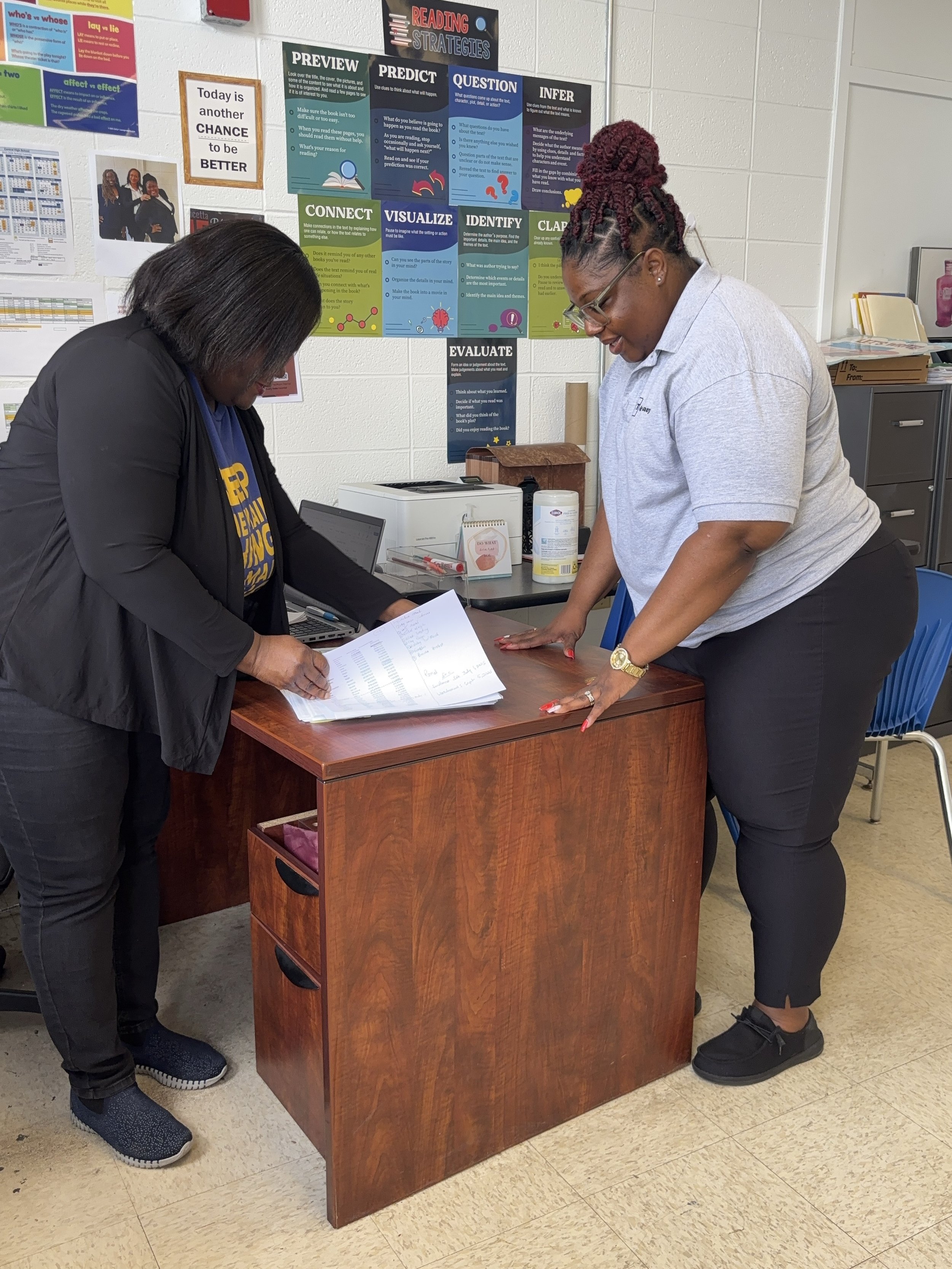 Two women are standing at a wooden desk, looking at papers. One woman, on the left, has black hair and is wearing a black jacket and black pants. The other woman, on the right, has braided reddish-purple hair, glasses, a light gray polo shirt, and black pants. The background includes educational posters on the wall, a printer, cleaning supplies, and a chair. The floor is beige tile.
