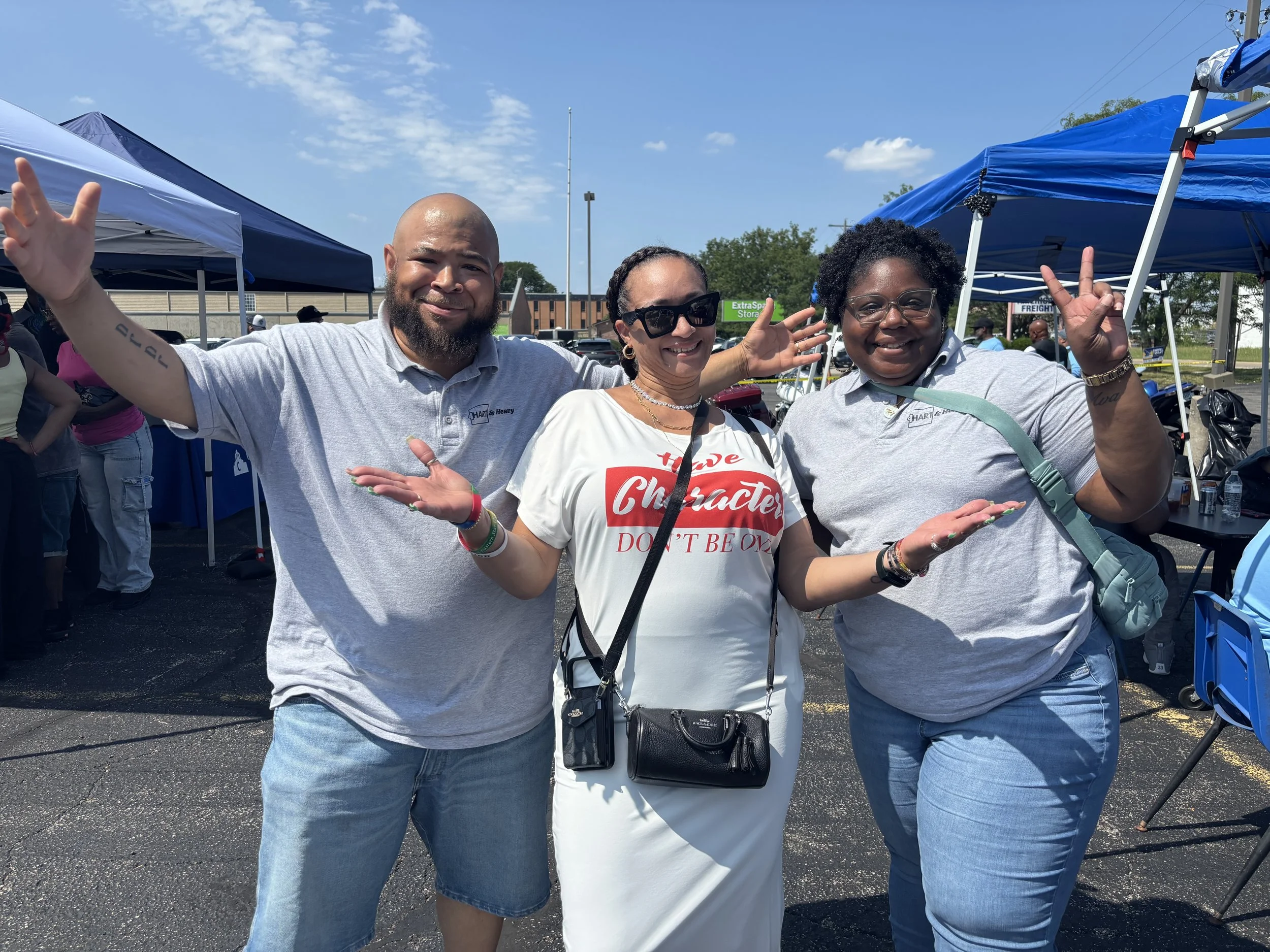 Three smiling people at an outdoor event, two women and one man, making peace signs with their hands. They are standing in front of blue tents with a parking lot and a building in the background.