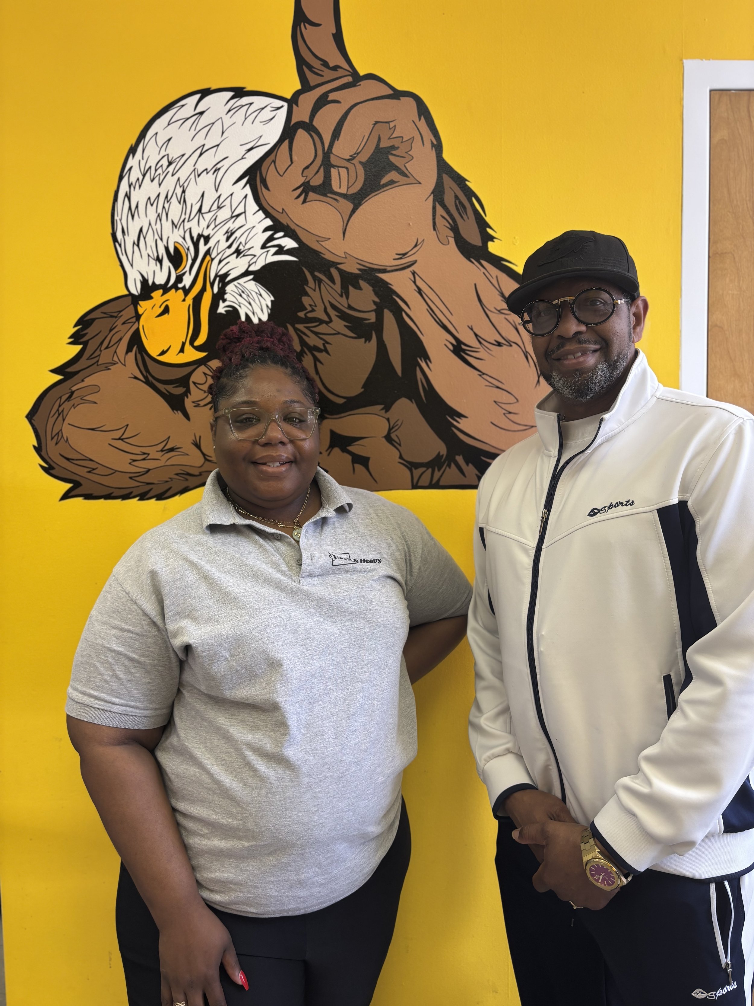 Two people standing in front of a yellow wall with a large cartoon-style illustration of a muscular eagle with white feathers, a yellow beak, and a brown body.
