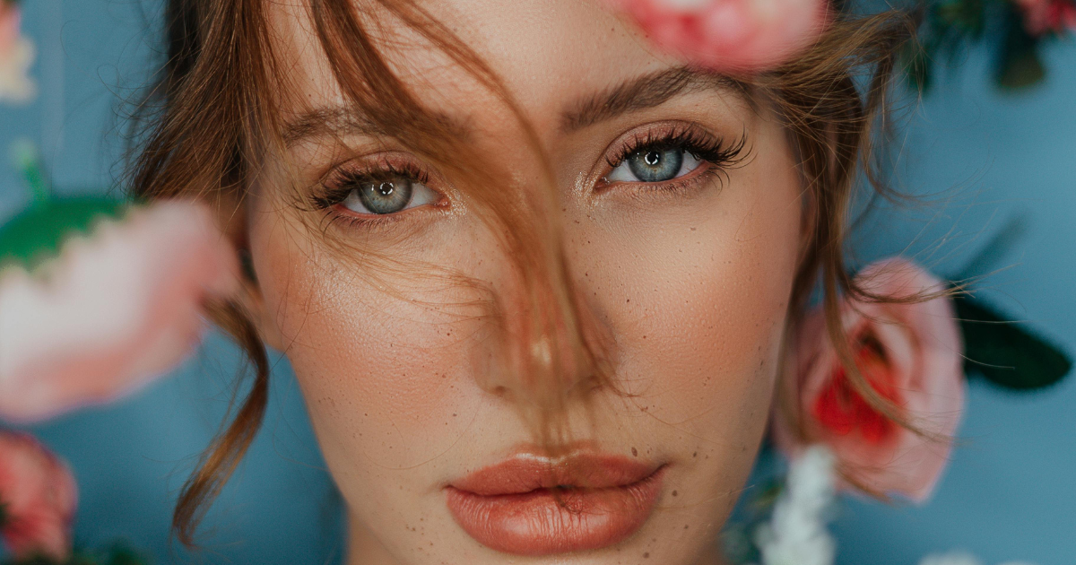 Close-up of a woman’s face surrounded by pink flowers, with blue eyes and makeup.
