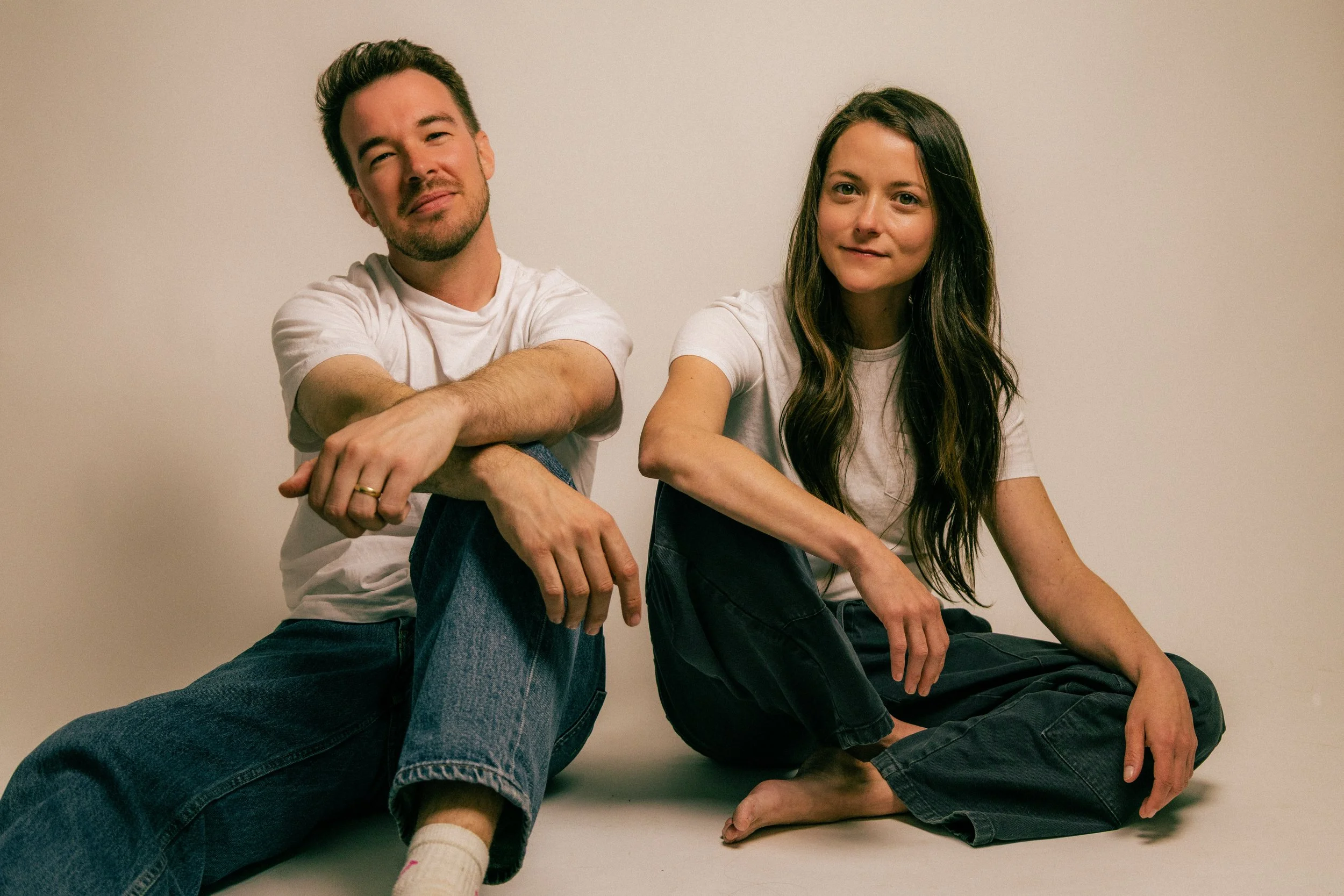 A young man and woman sitting on the floor against a plain wall. The man is wearing a white t-shirt and jeans, and has his arms crossed over his knees. The woman is in a white t-shirt and dark pants, with long brown hair, and is barefoot. Both are looking at the camera with relaxed expressions.
