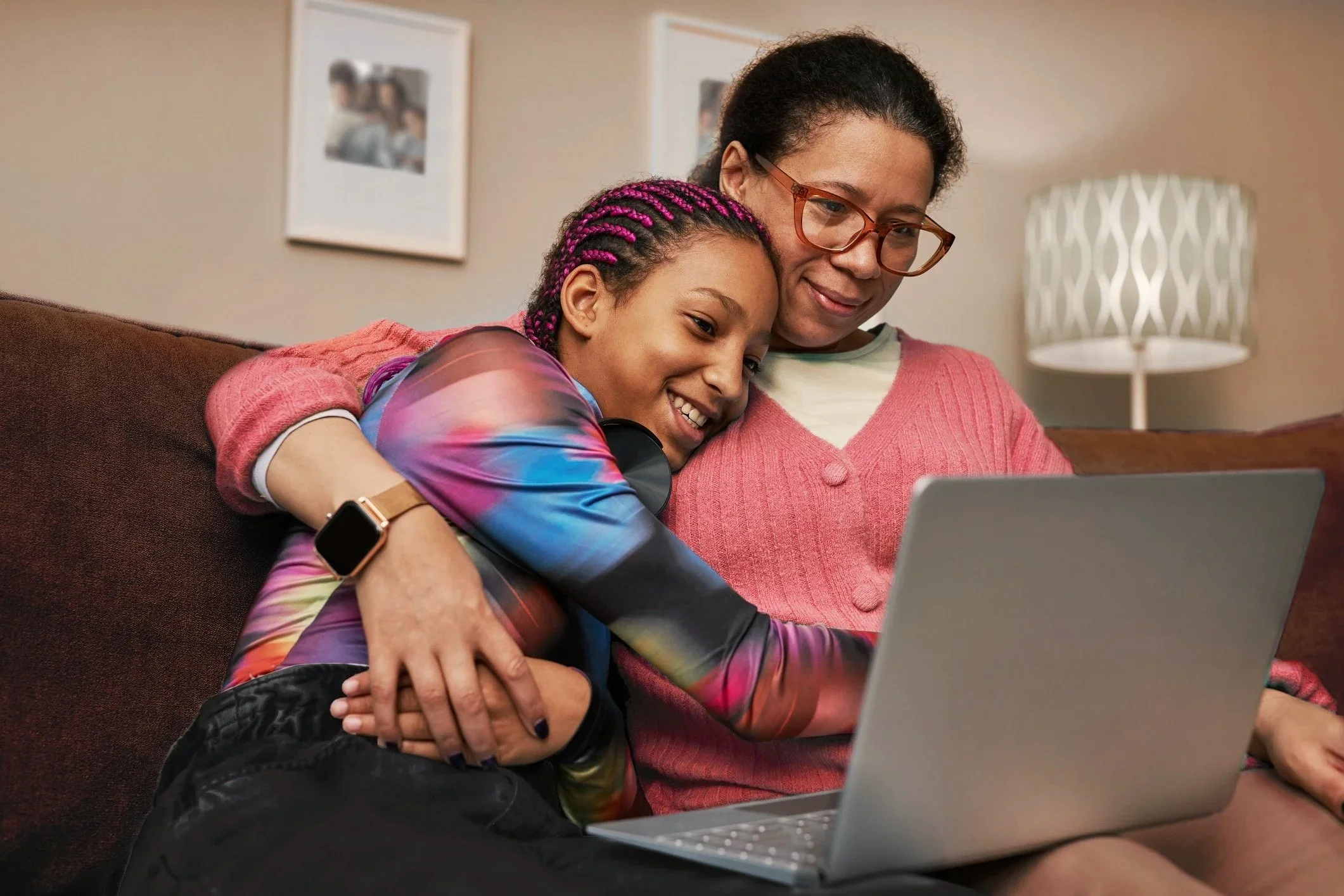 A mom and teen are sitting close together on a couch so they can share the laptop while video chatting. The mom has her arm tossed around her kid, and the teen is smiling with teeth showing.