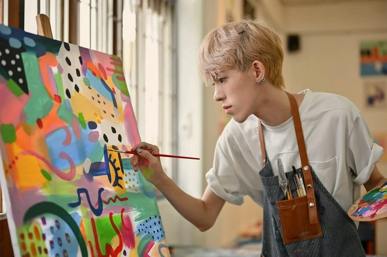 A teen wearing a tshirt and an apron is painting a colorful graphic canvas. Their hair is bleached and they have multiple piercings.