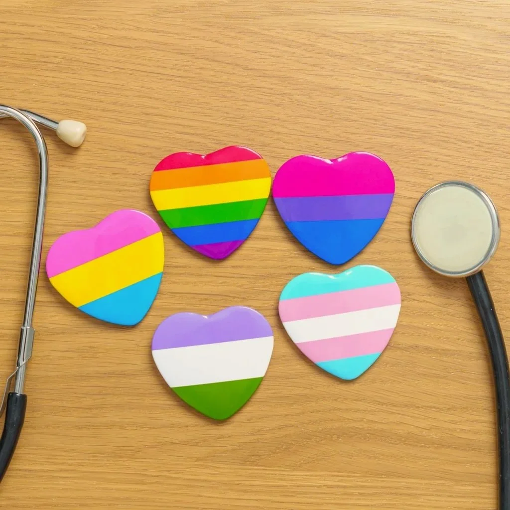 Heart-shaped pins with stripes representing flags: Pride, Transgender, Bisexual, Pansexual, Genderqueer flags. To the right and left of these hearts is a stethoscope earpiece and and diaphragm (aka the chest piece - what is used to listen).