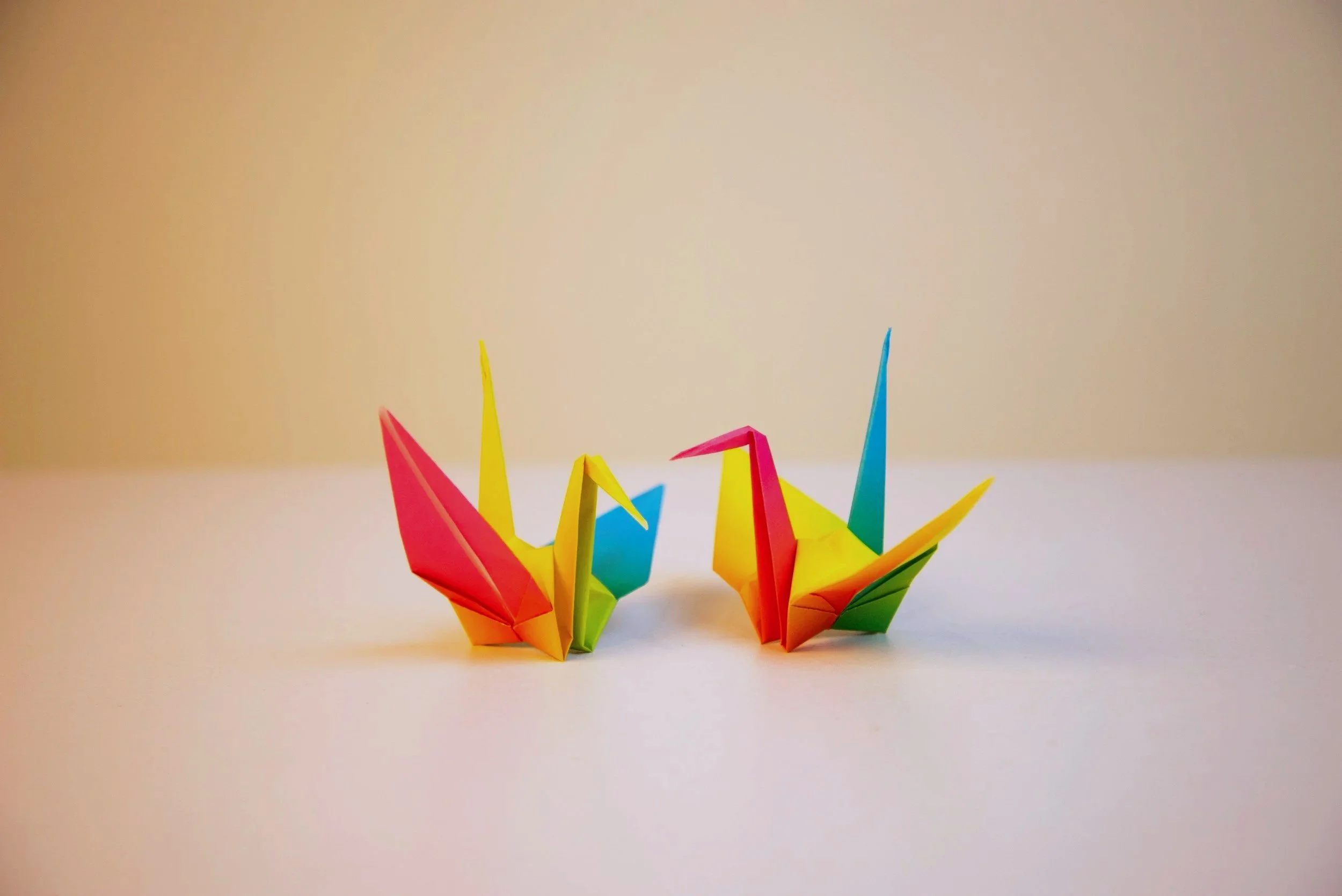Two rainbow ombre origami paper cranes sit on a table top.