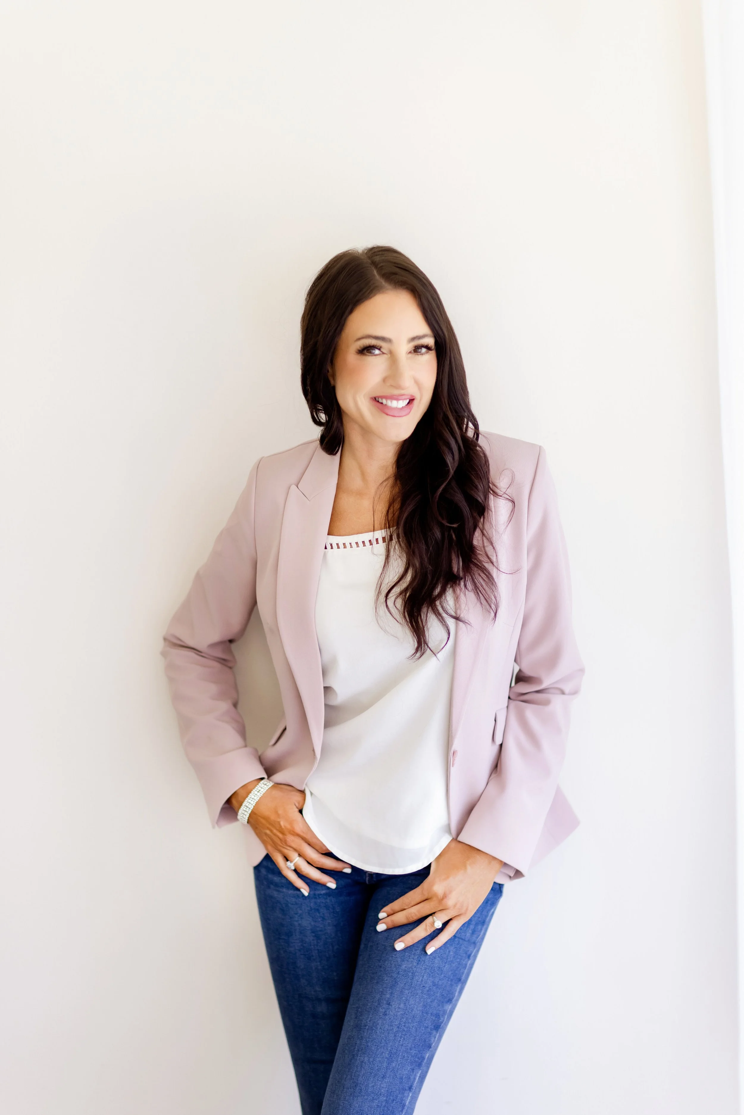 A woman with long dark hair wearing a light pink blazer, white top, and blue jeans, standing against a plain white wall and smiling.