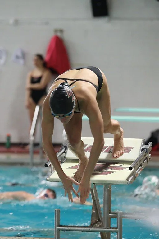 Swimmer on starting block preparing to dive into the pool at an indoor swimming competition.