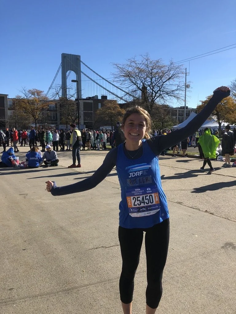 Young woman smiling and celebrating at a marathon event near a bridge, wearing a blue marathon bib and black leggings.