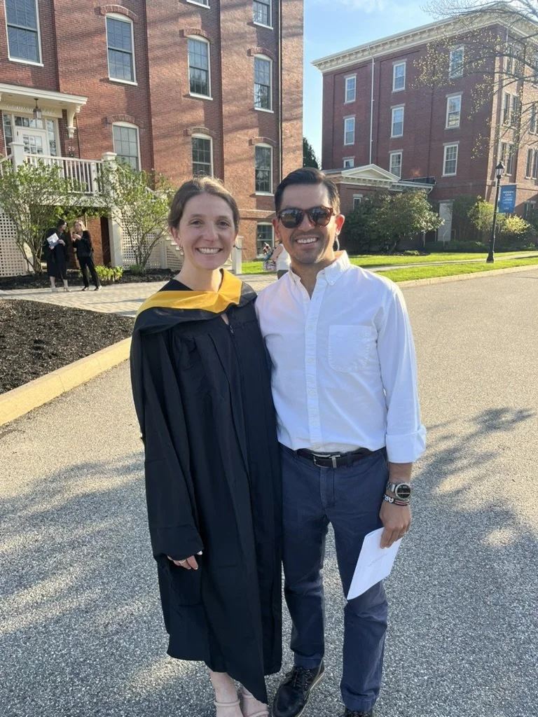 A woman in a graduation gown and stole standing next to a man in a white shirt and dark pants, both smiling outdoors near brick buildings and trees.