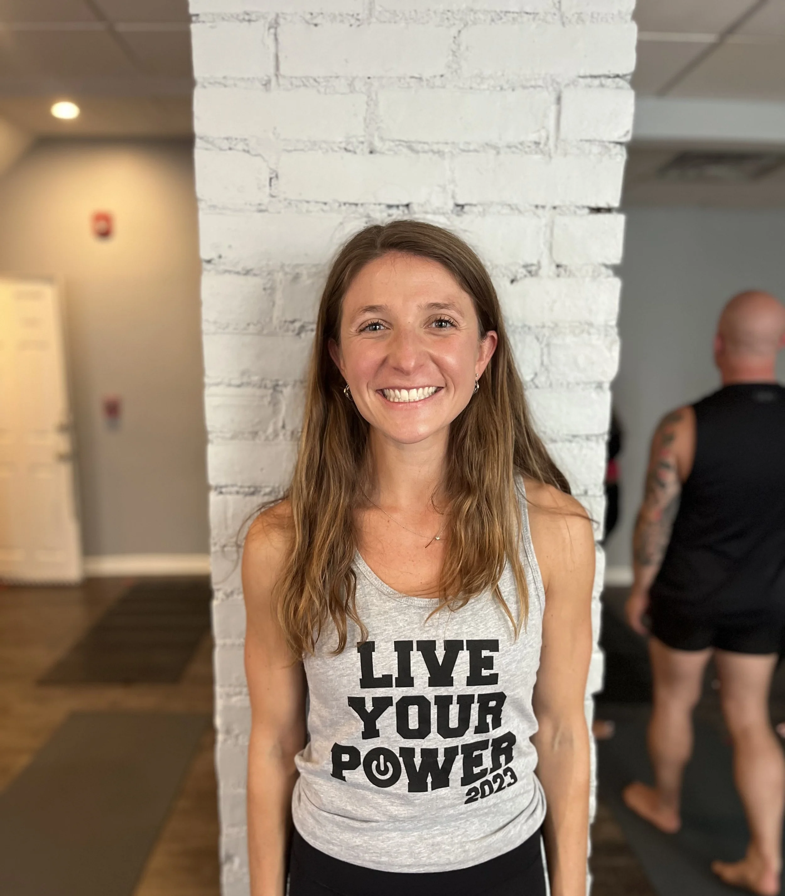 Close-up of a smiling woman with long brown hair standing in front of a white brick wall, wearing a tank top with the words 'LIVE THE POWER 2022' printed on it. A person with tattoos wearing a black sleeveless shirt is visible in the background.