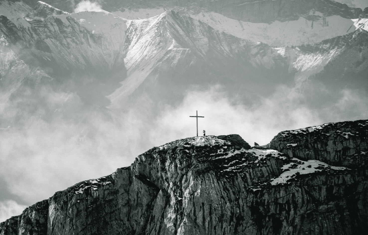 A mountain landscape in black and white with a cross on a rocky summit in the foreground and snow-capped mountains in the background, with clouds and mist surrounding the peaks.